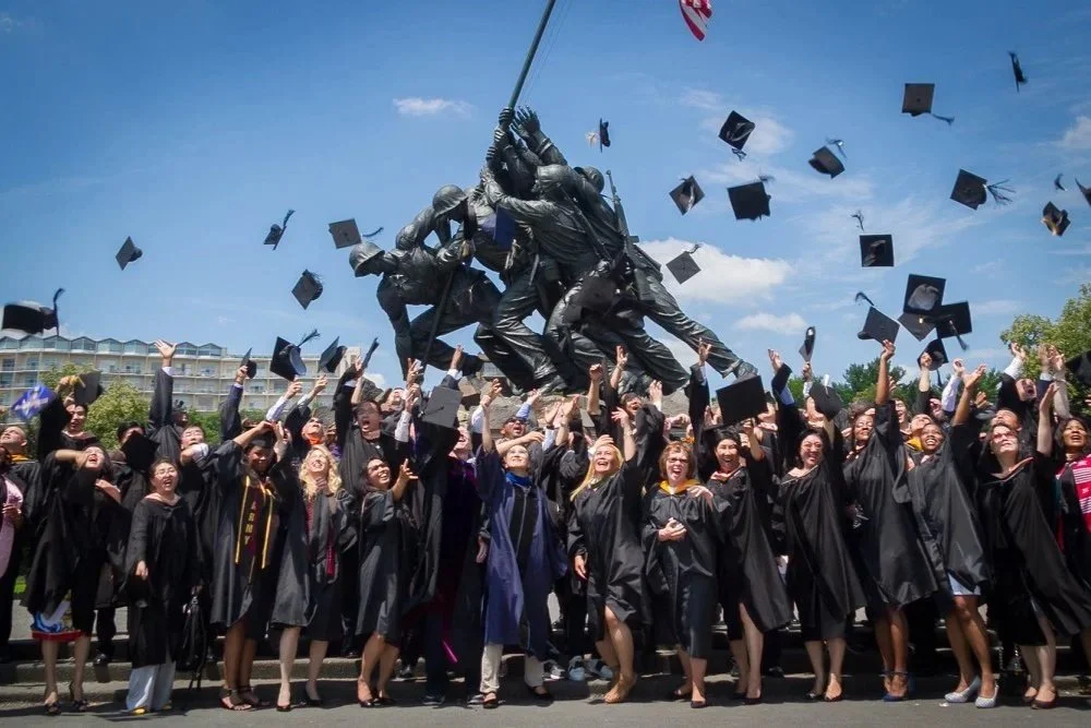 Graduates in black gowns celebrating and throwing caps in the air at a graduation ceremony outside, with a large monument in the background and blue sky.