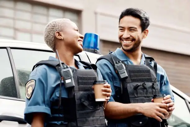 Two smiling police officers, one Black woman and one man, standing outside a police vehicle, holding coffee cups and sharing a light moment.