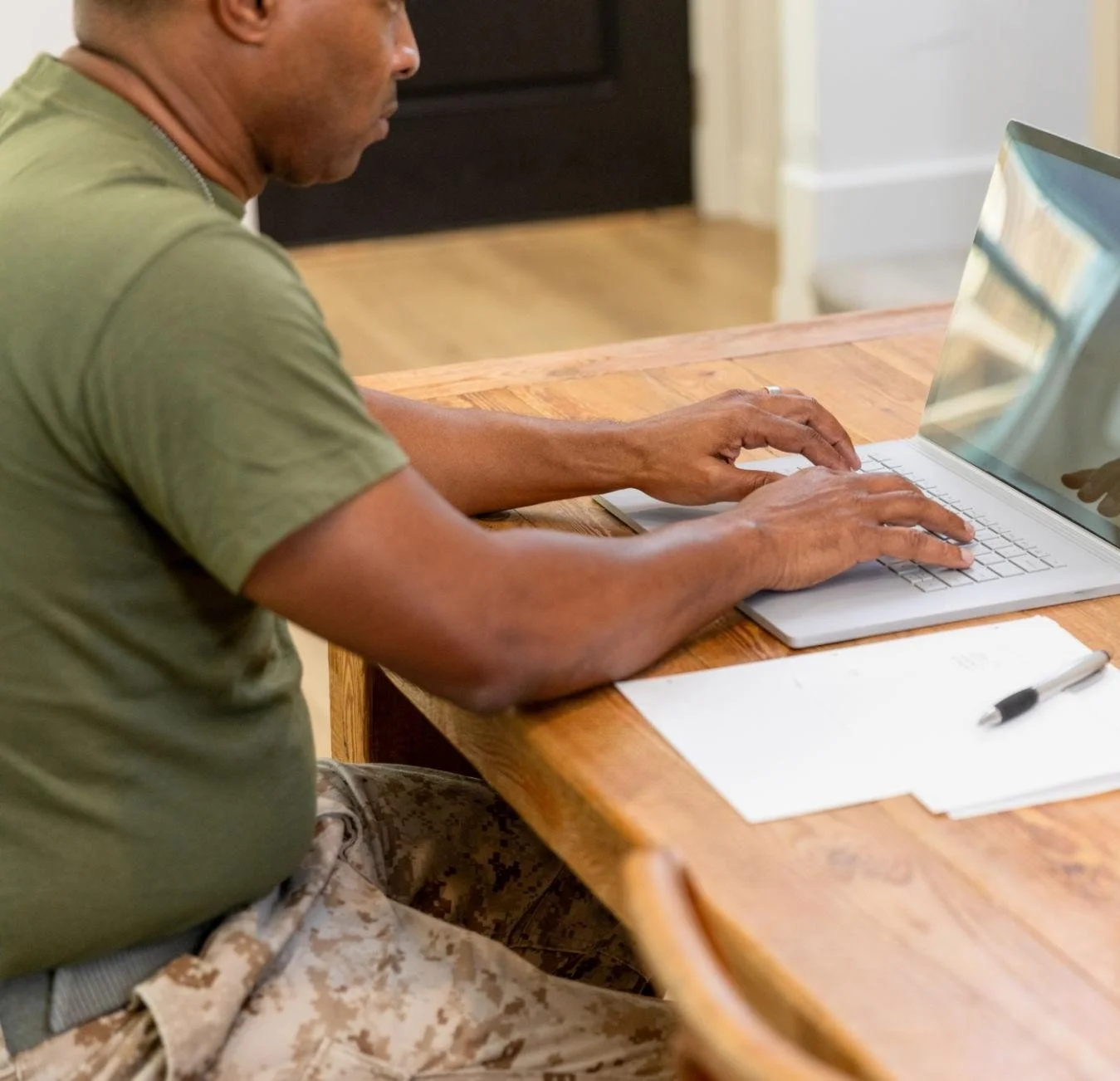 A man in military camouflage pants and a green t-shirt working on a laptop at a wooden table with a white paper and a black pen.