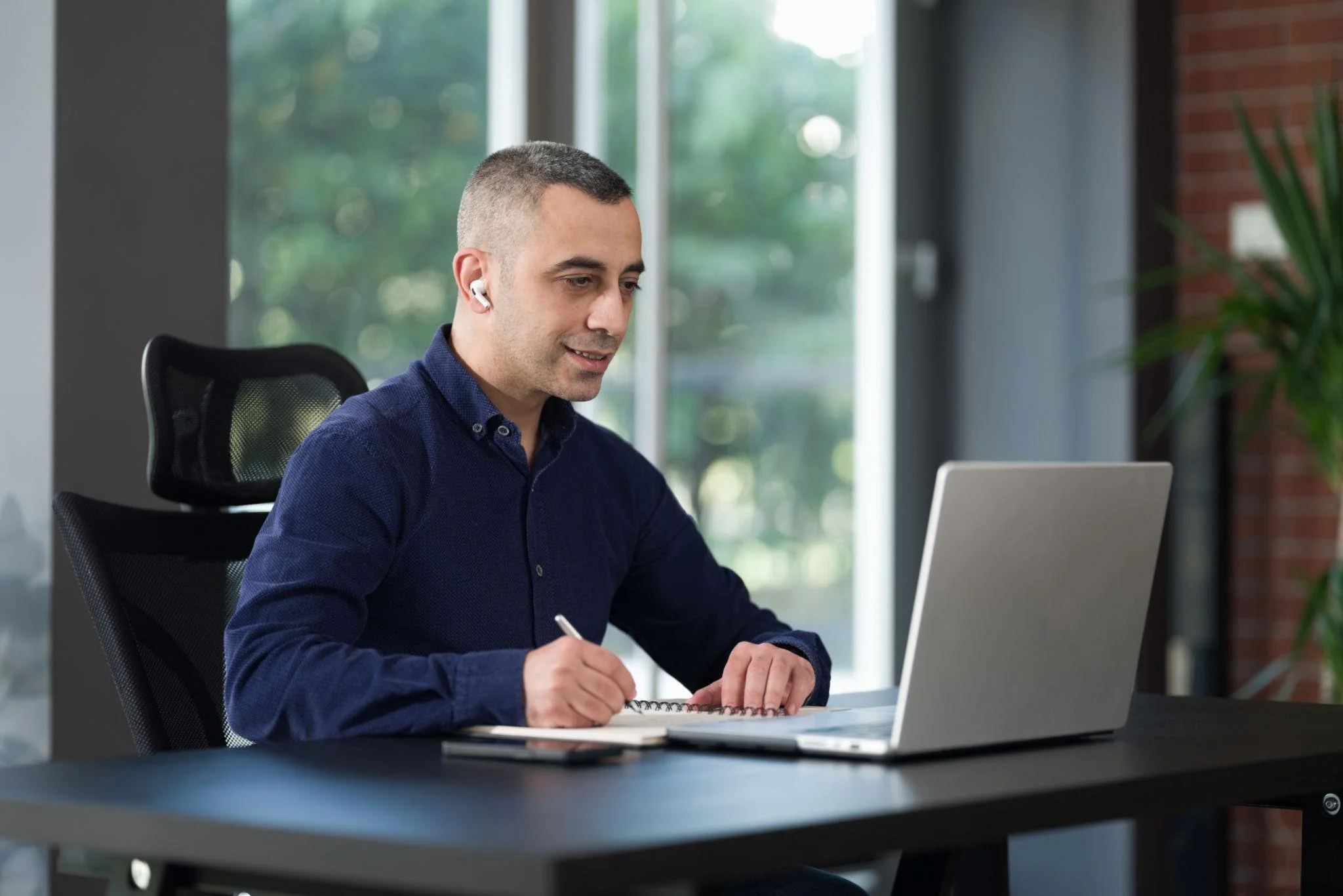 A man sitting at a desk with a laptop, taking notes with a pen, wearing wireless earbuds, in a modern office with large windows and greenery outside.