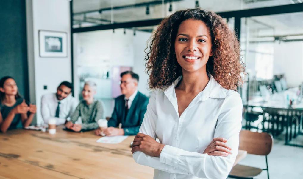 A confident woman with curly hair standing in front of a group of diverse professionals in a modern office conference room.
