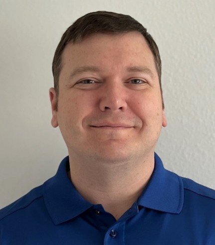 A man with short brown hair and a slight smile, wearing a blue collared shirt, standing against a white wall.