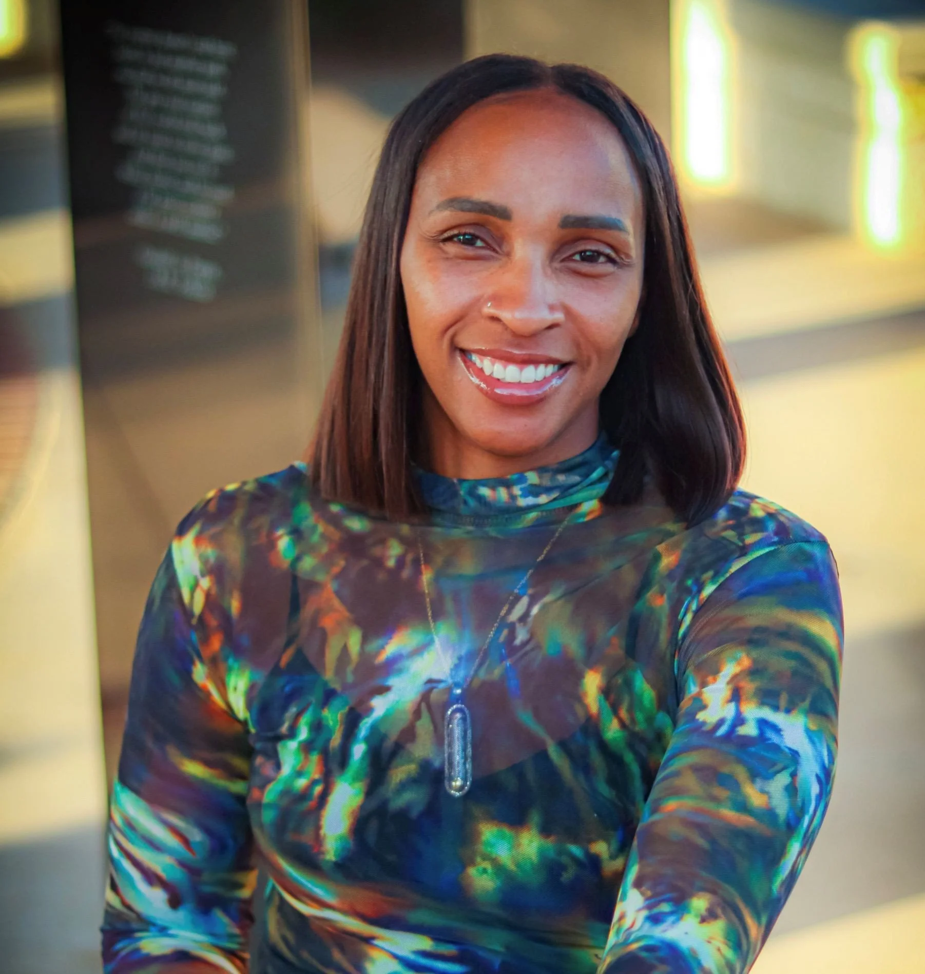 A smiling woman with shoulder-length dark hair stands in front of a blurred background of neon lights, wearing a colorful, abstract patterned long-sleeve top and a necklace with a vertical pendant.