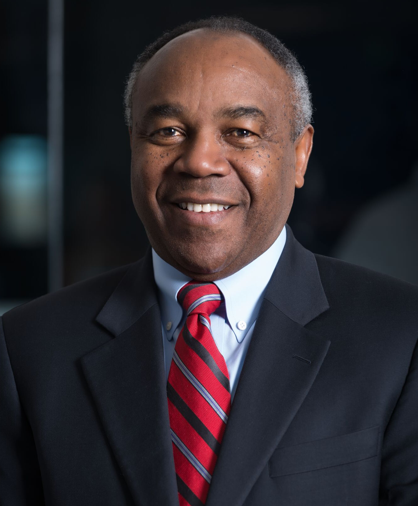 A man wearing a dark suit, white shirt, and a red striped tie, smiling and looking at the camera.