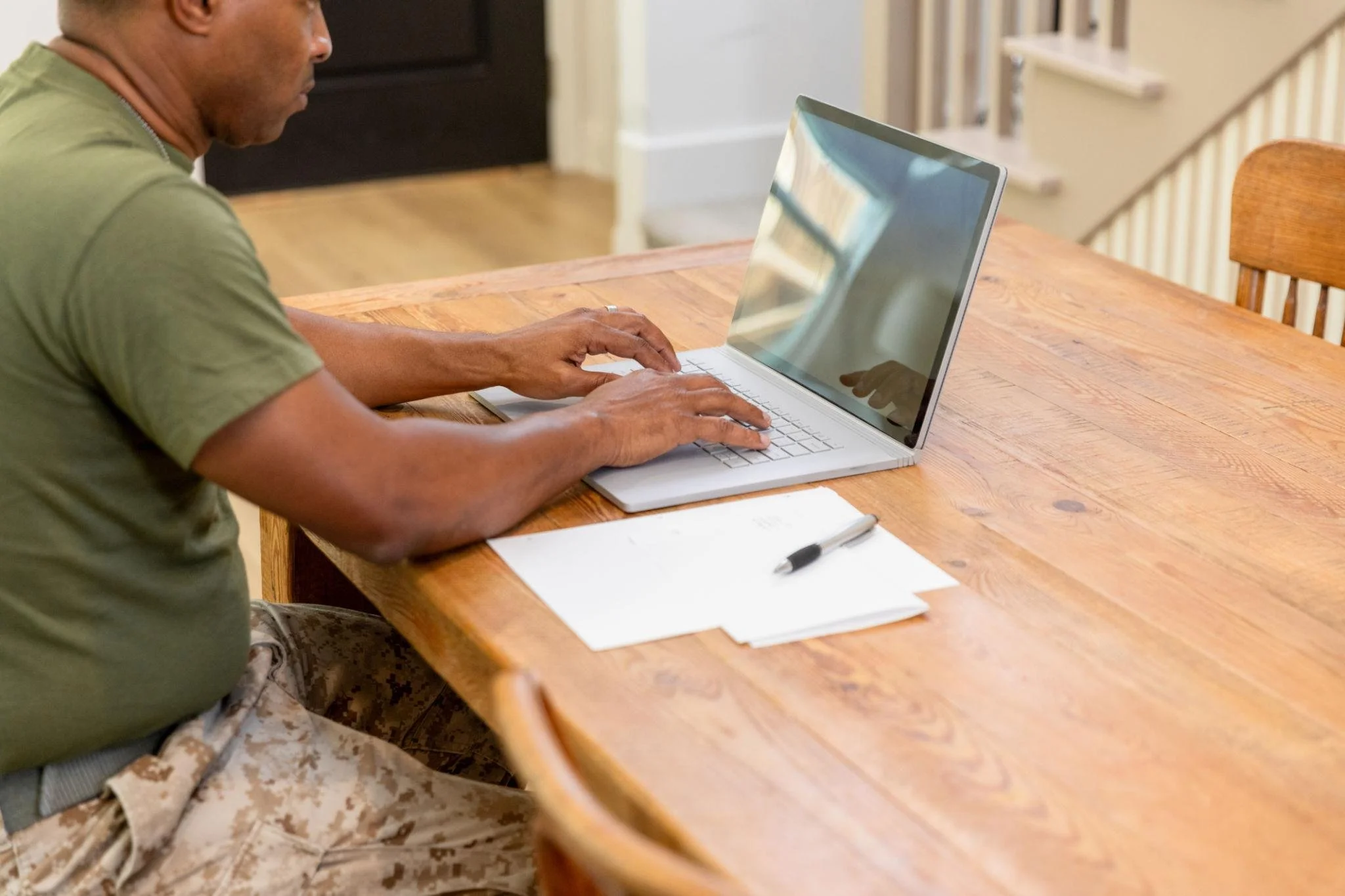 Man working on a silver laptop at a wooden table with papers and a pen, in a home setting.