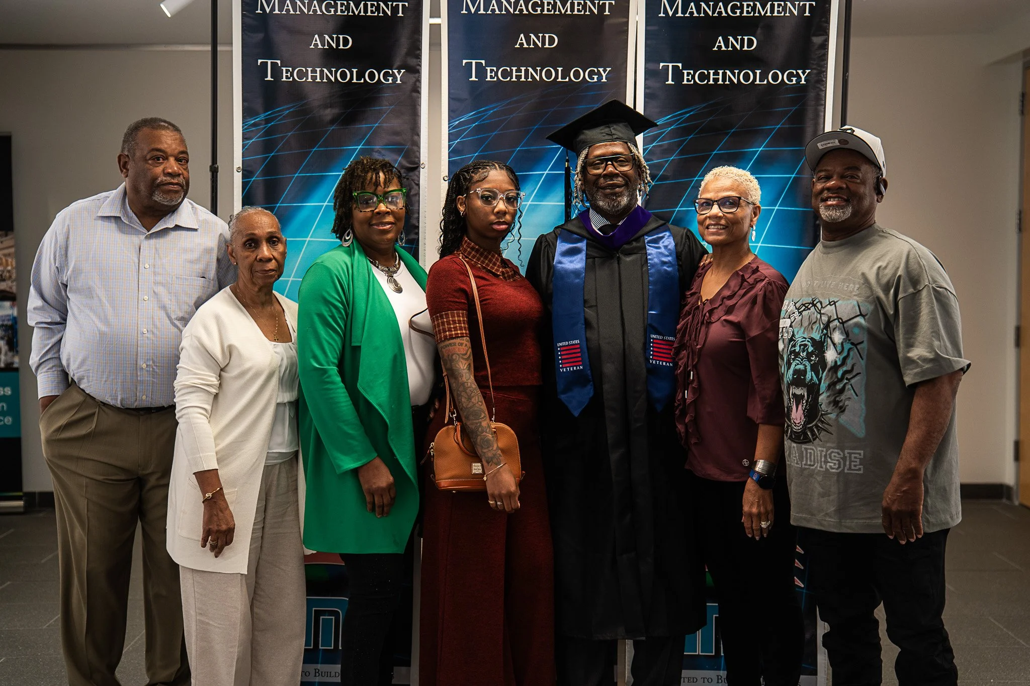 Group of eight people posing at a graduation ceremony, with a graduate in cap and gown in the center. The background has banners reading 'Management and Technology.'