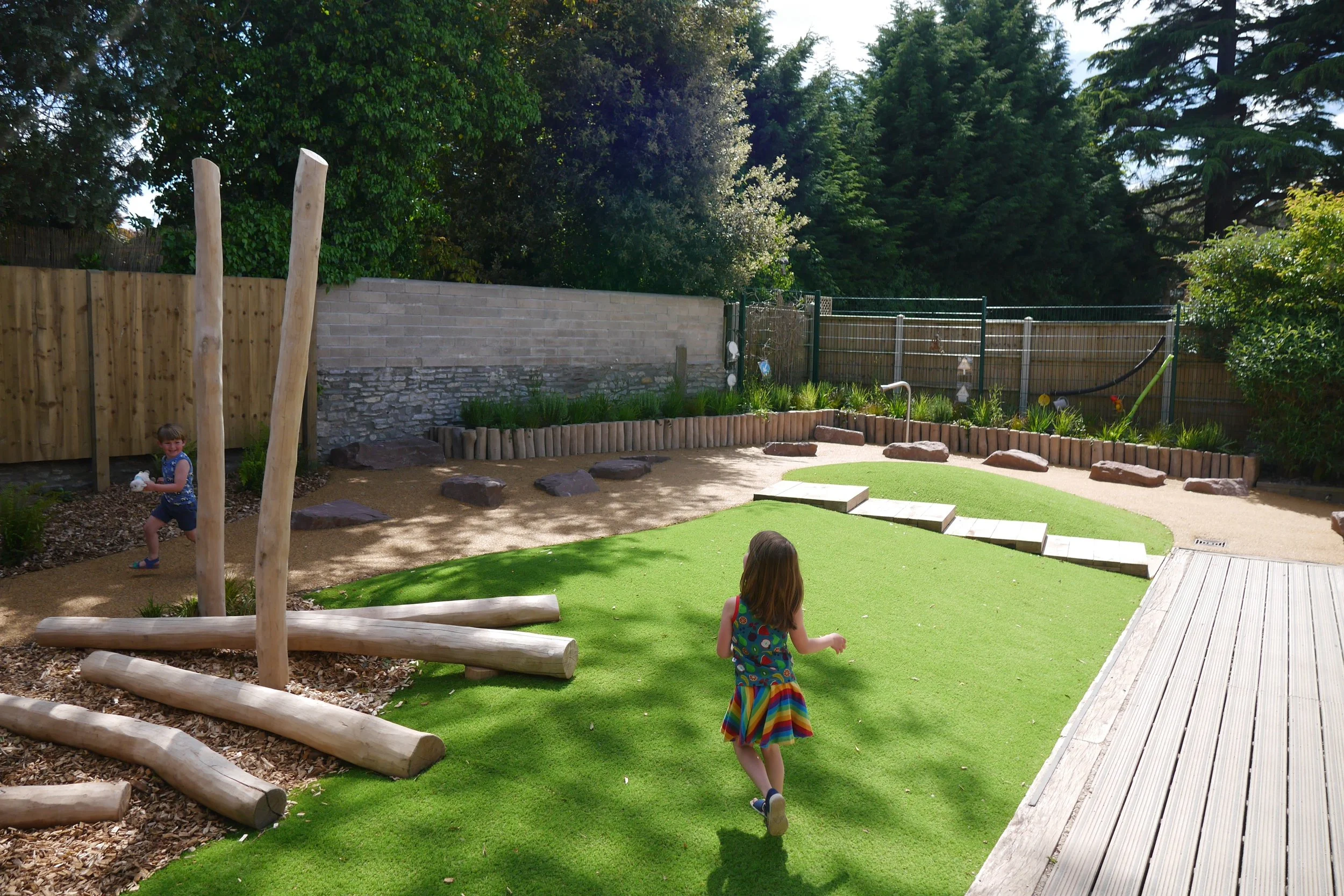 Children playing in a backyard with artificial grass, wooden logs, rocks, and a garden with shrubs and trees, enclosed by a fence.