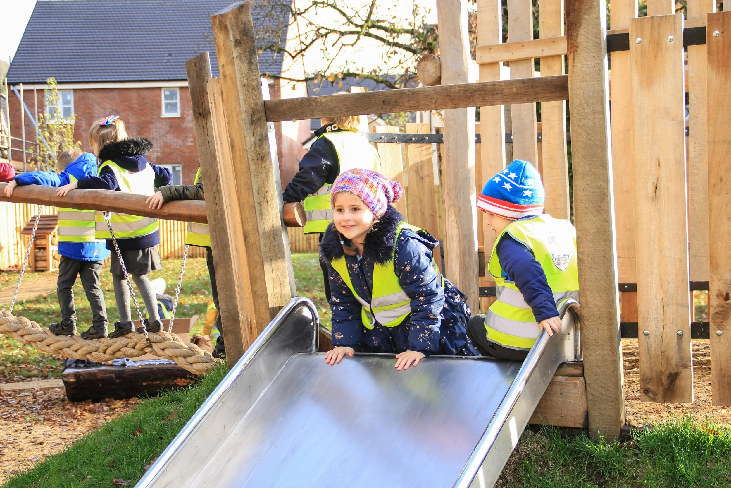 Children playing on a playground slide and climbing on a wooden structure, wearing yellow safety vests and winter hats.