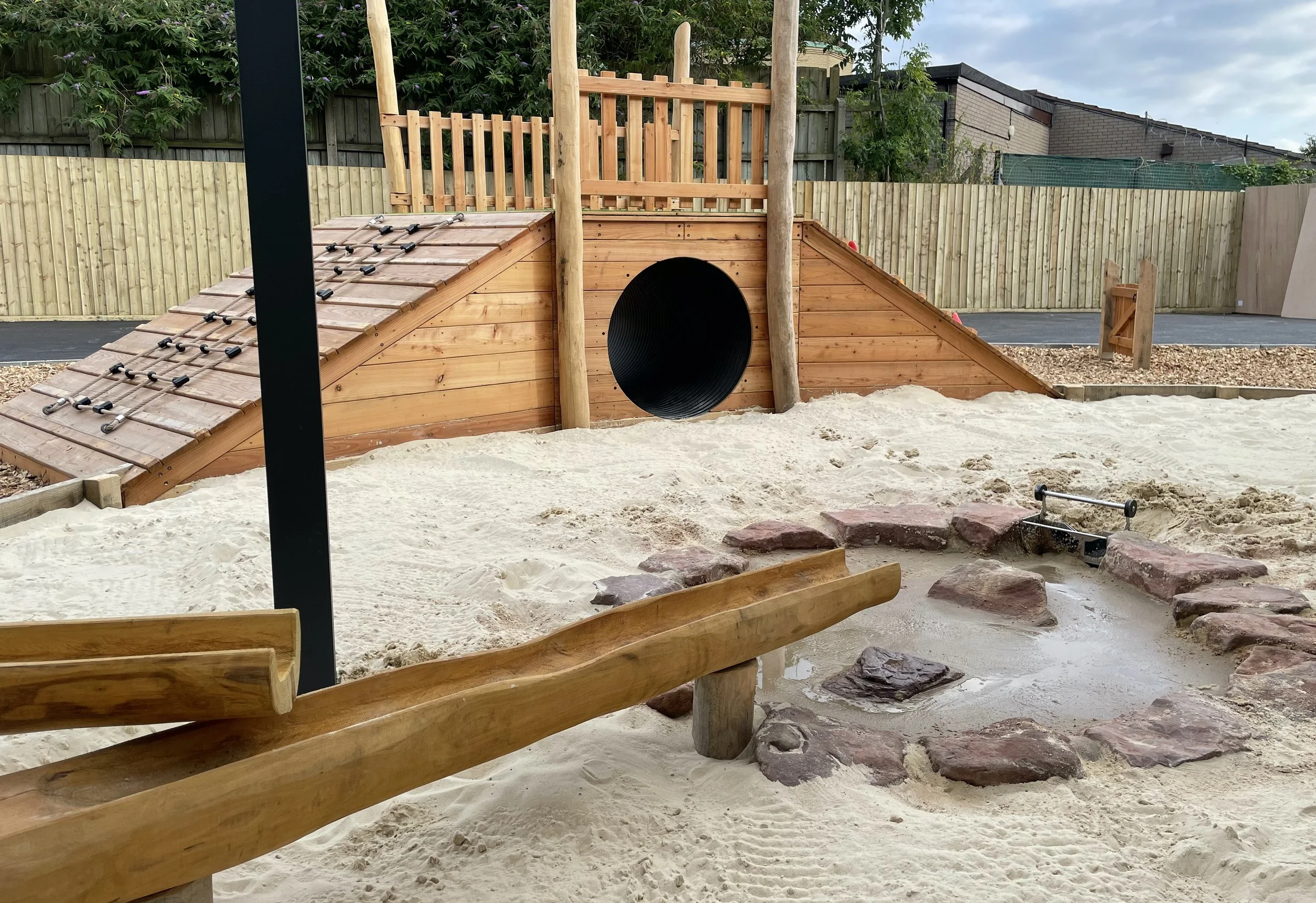 A children's playground featuring a wooden slide with climbing rungs, a tunnel entrance, and a sandbox with a small water feature surrounded by rocks.