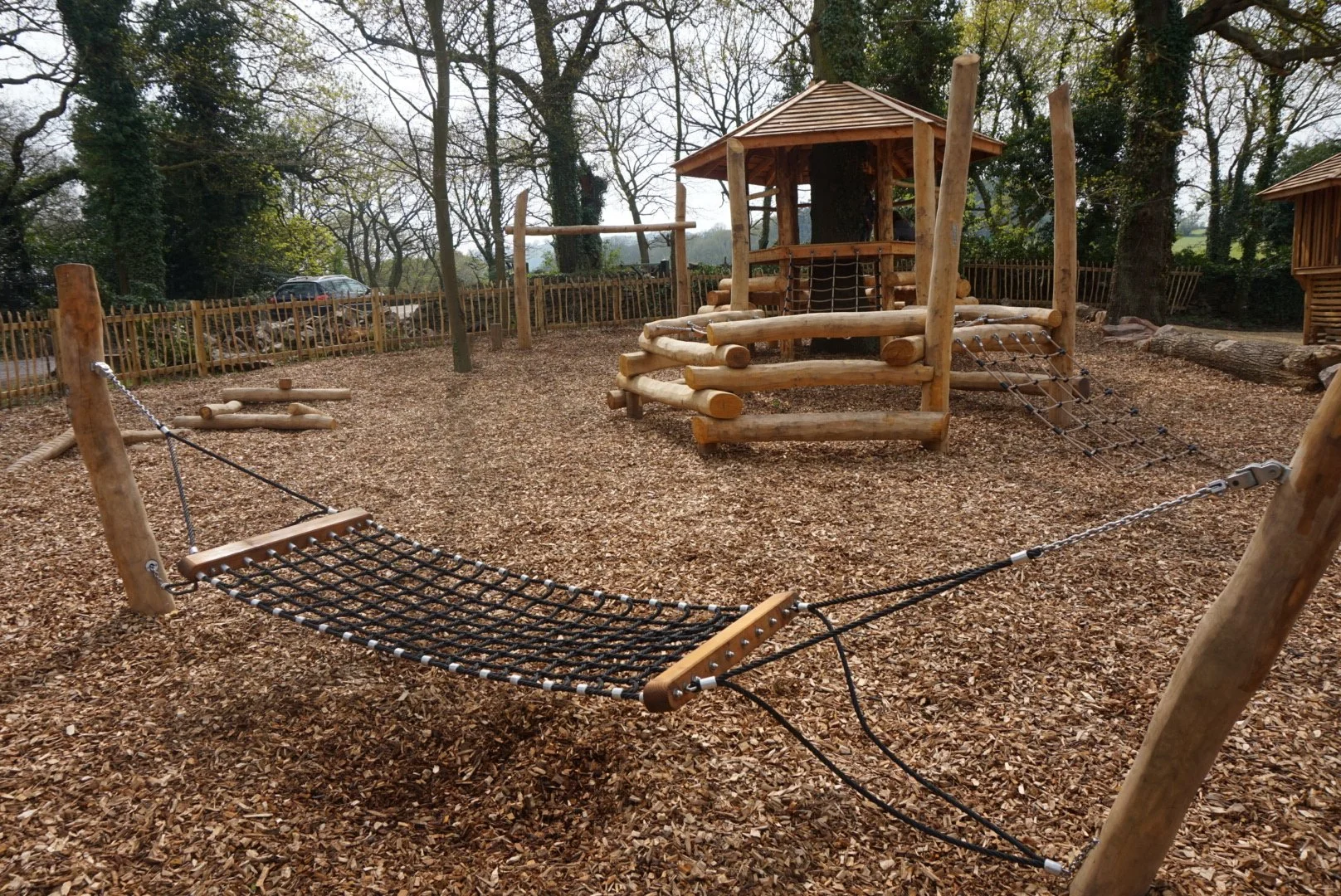 Wooden playground structure with a bridge, ropes, and a small shelter, surrounded by children-friendly wood chips and trees.