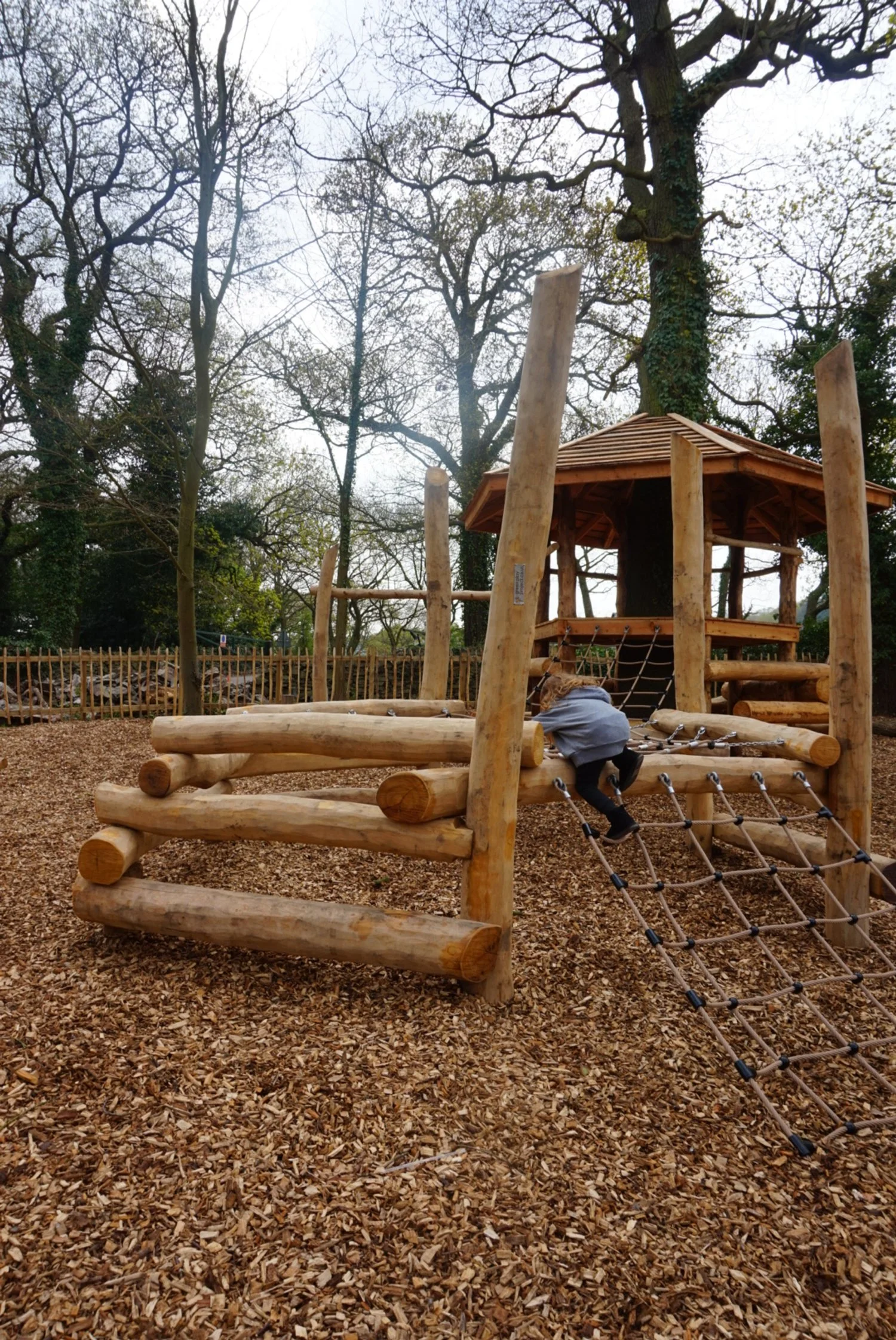 Child climbing a wooden playground structure with climbing ropes in a forested park.