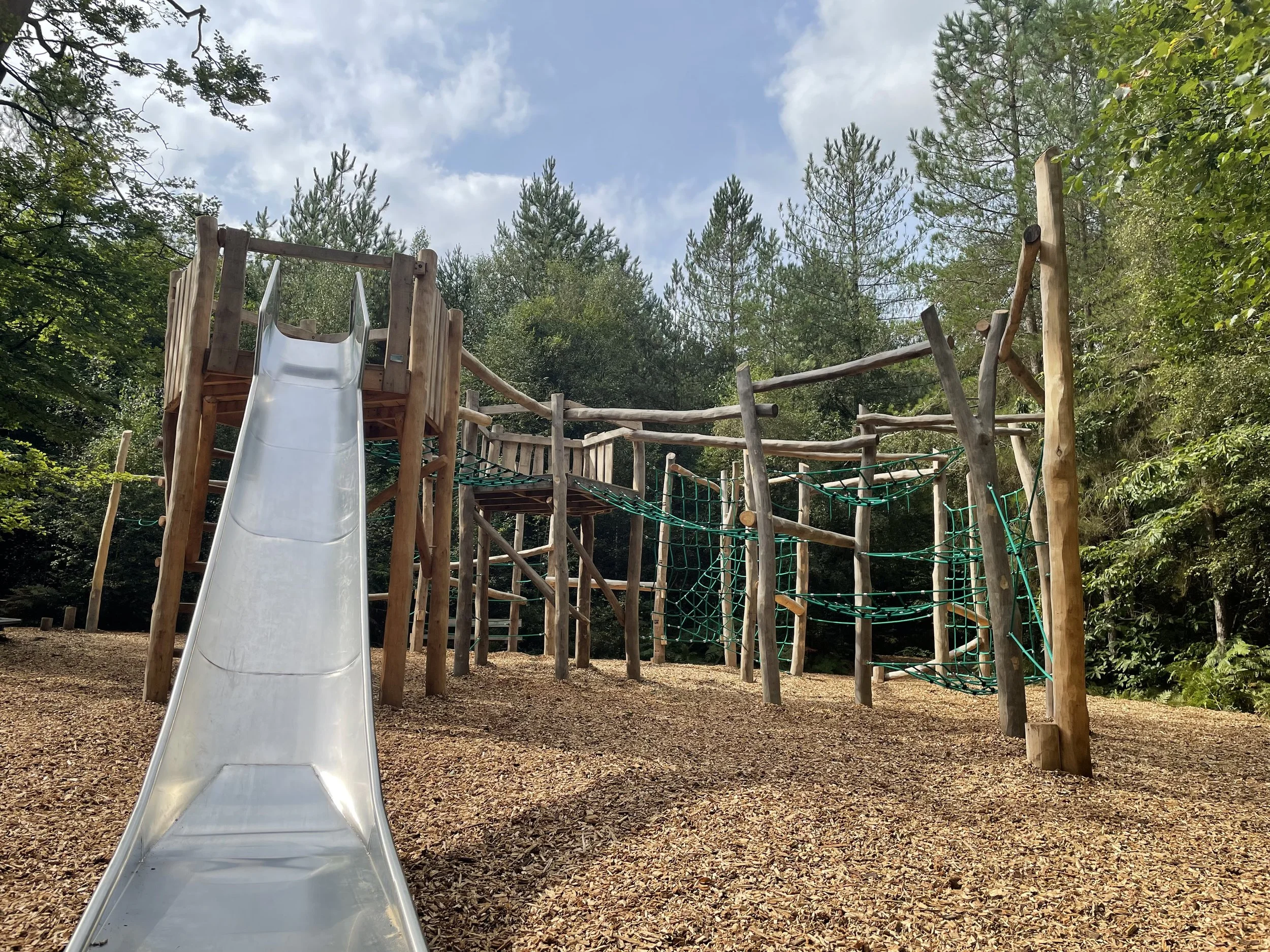 Outdoor wooden playground structure with a metal slide, rope bridges, and climbing nets surrounded by trees and mulch ground.