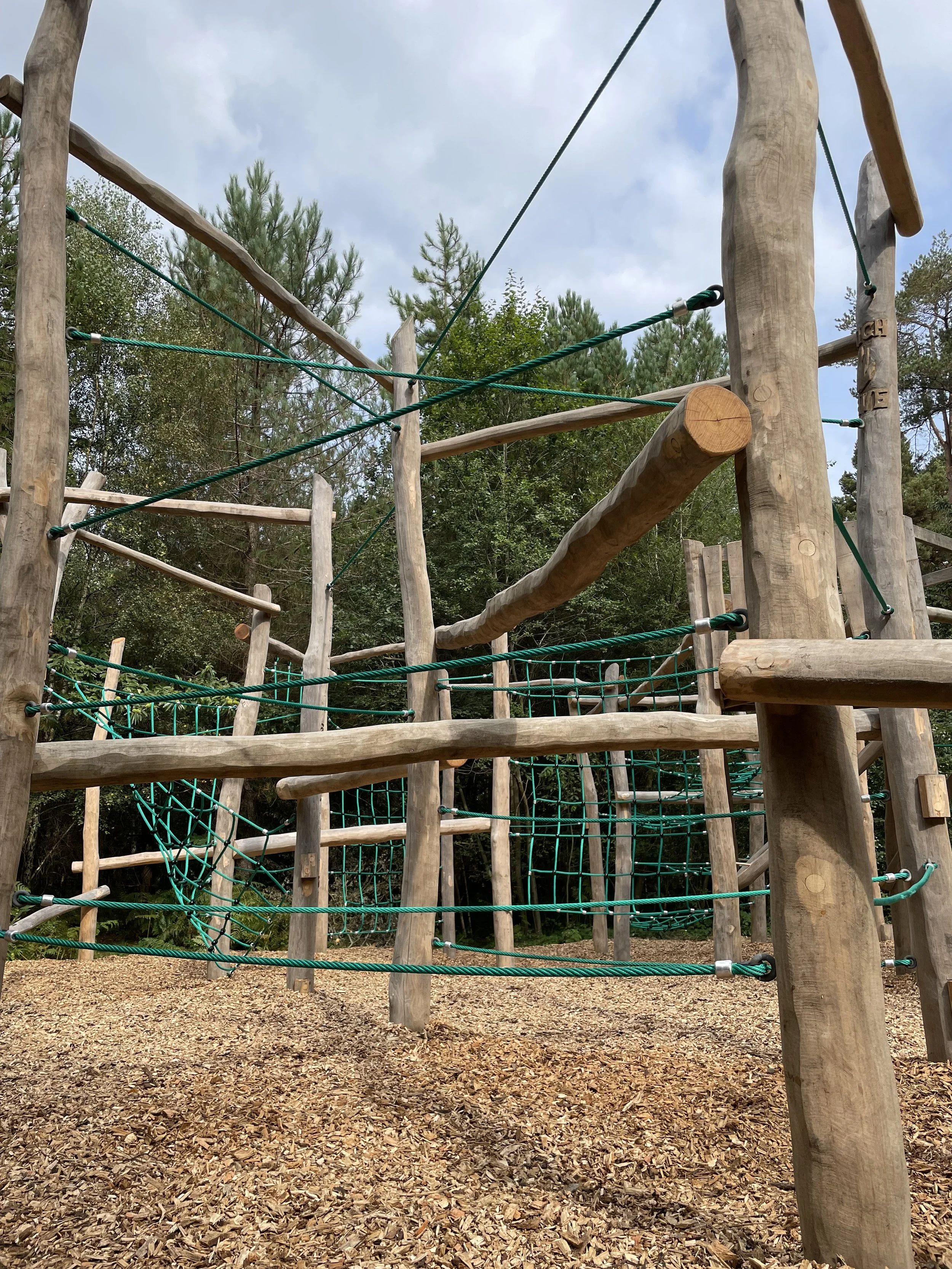 Wooden climbing structure with ropes and logs in a playground, surrounded by trees and wood chips.