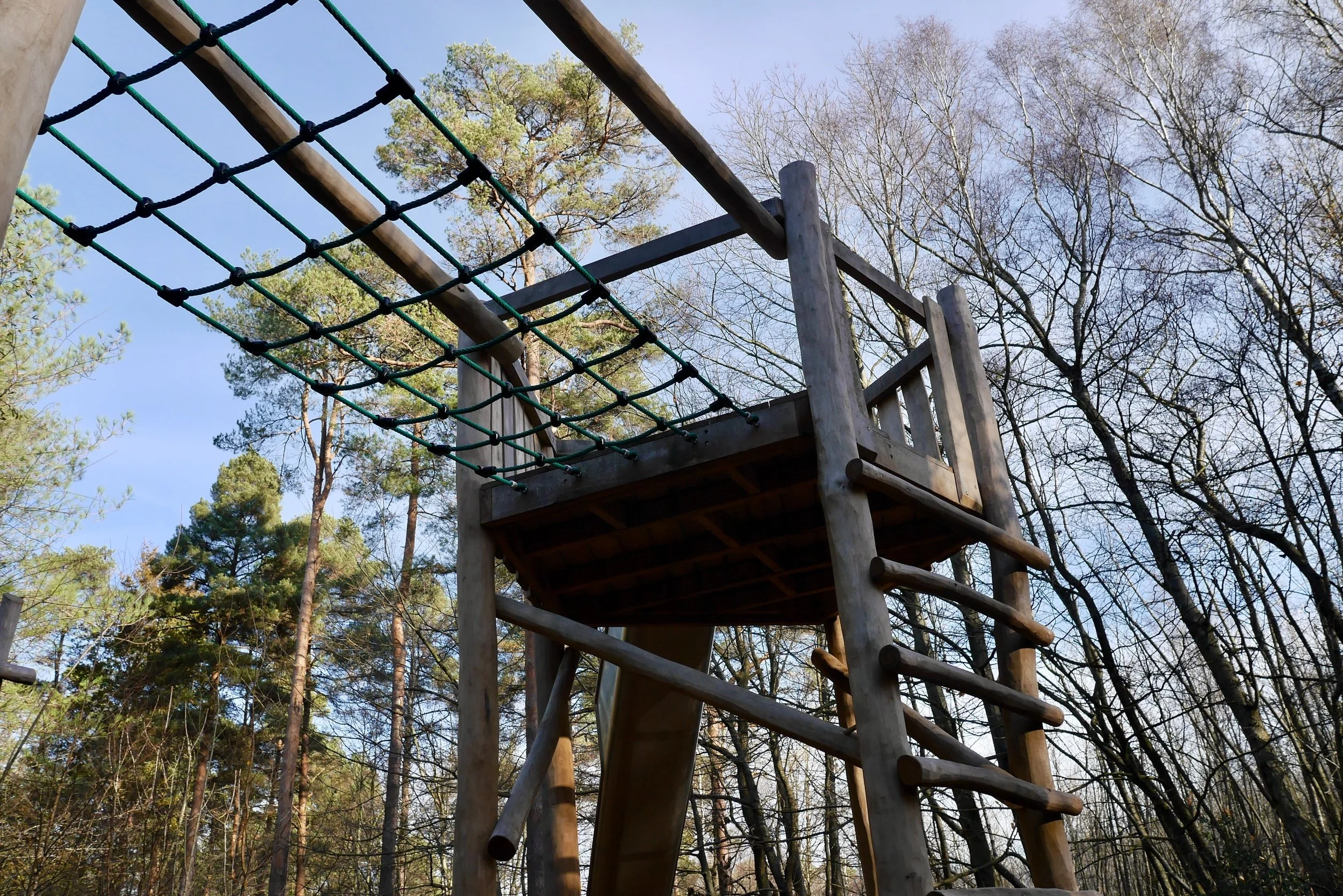 Wooden playground structure with a net climbing area and slide, surrounded by trees with no leaves, under a clear blue sky.