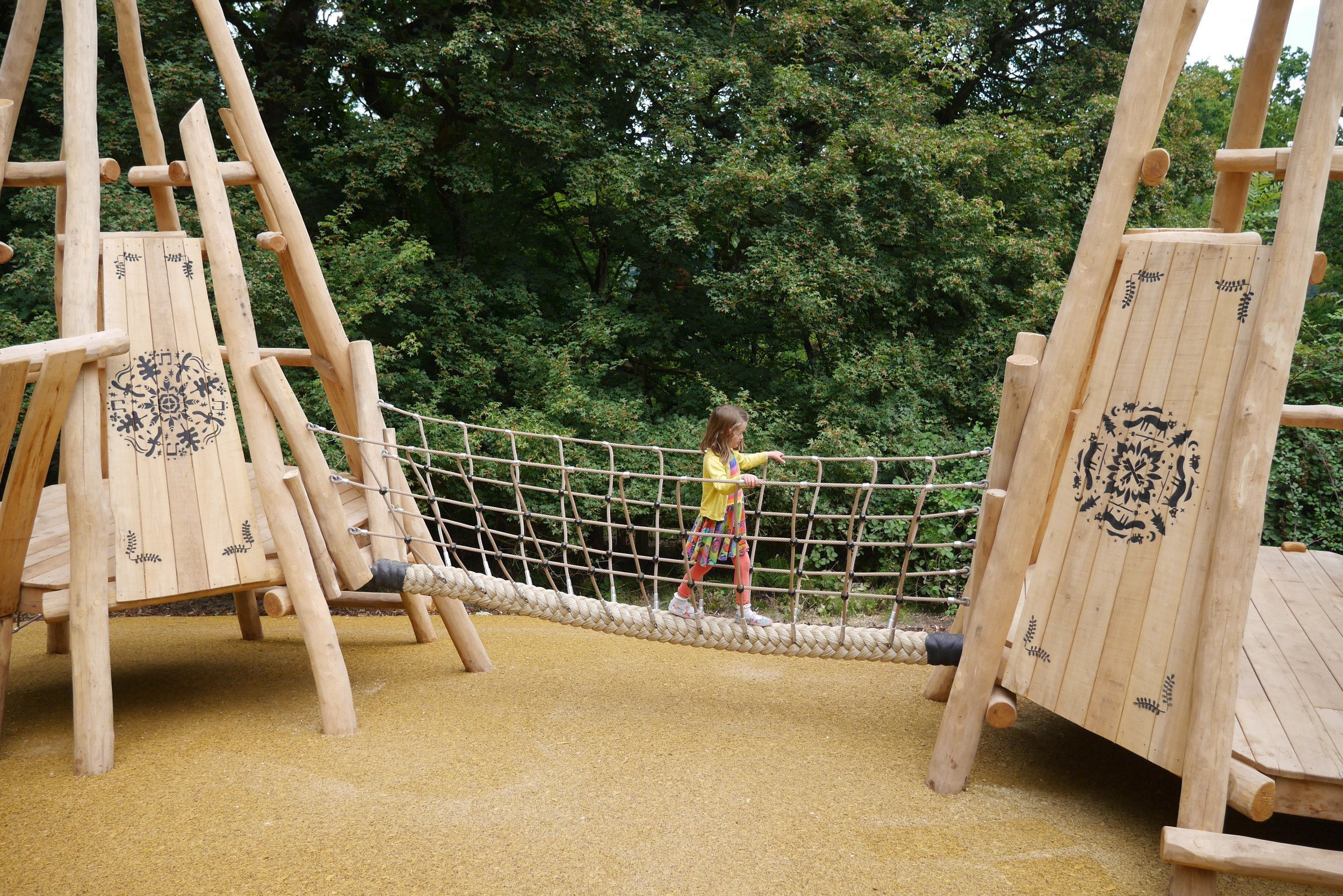 A young girl walking on a rope bridge in a playground with wooden structures and green trees in the background.