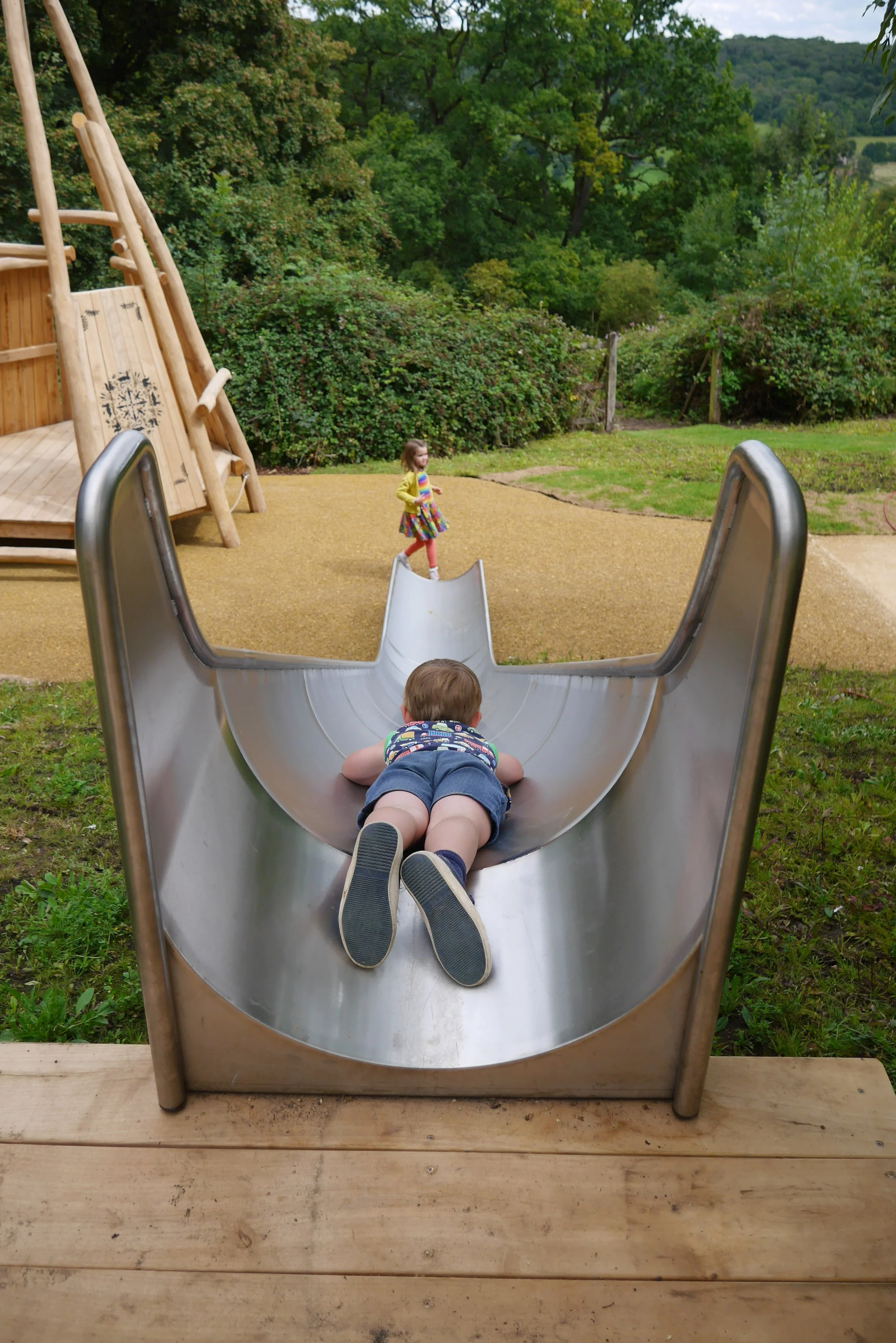 Child crawling down a metal slide at a playground, with a girl standing in the background on a dirt surface surrounded by green trees.