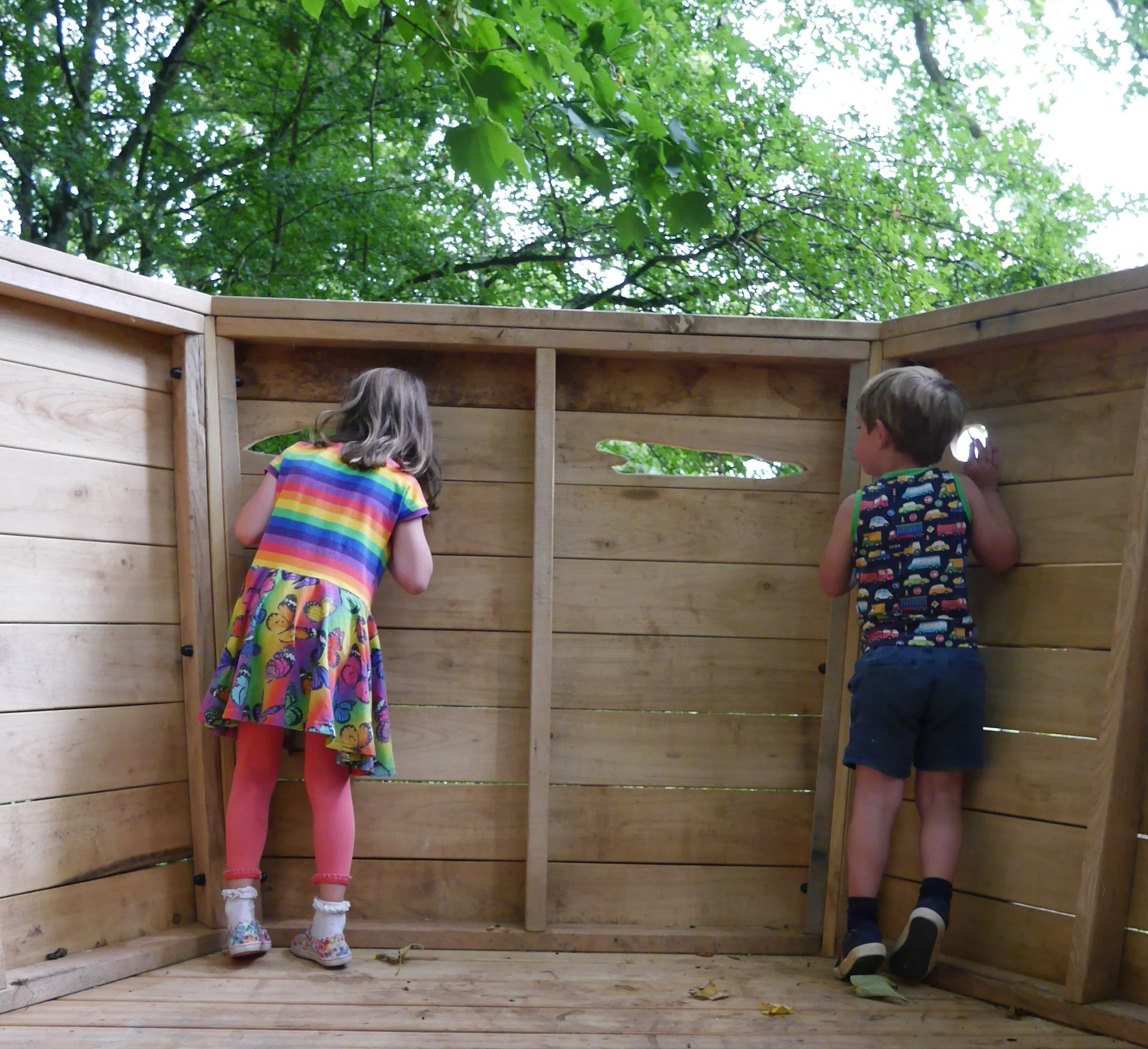 Two children, a girl in a colorful dress and pink tights, and a boy in a sleeveless shirt with vehicle patterns and shorts, are peering through holes in a wooden outdoor play fort.