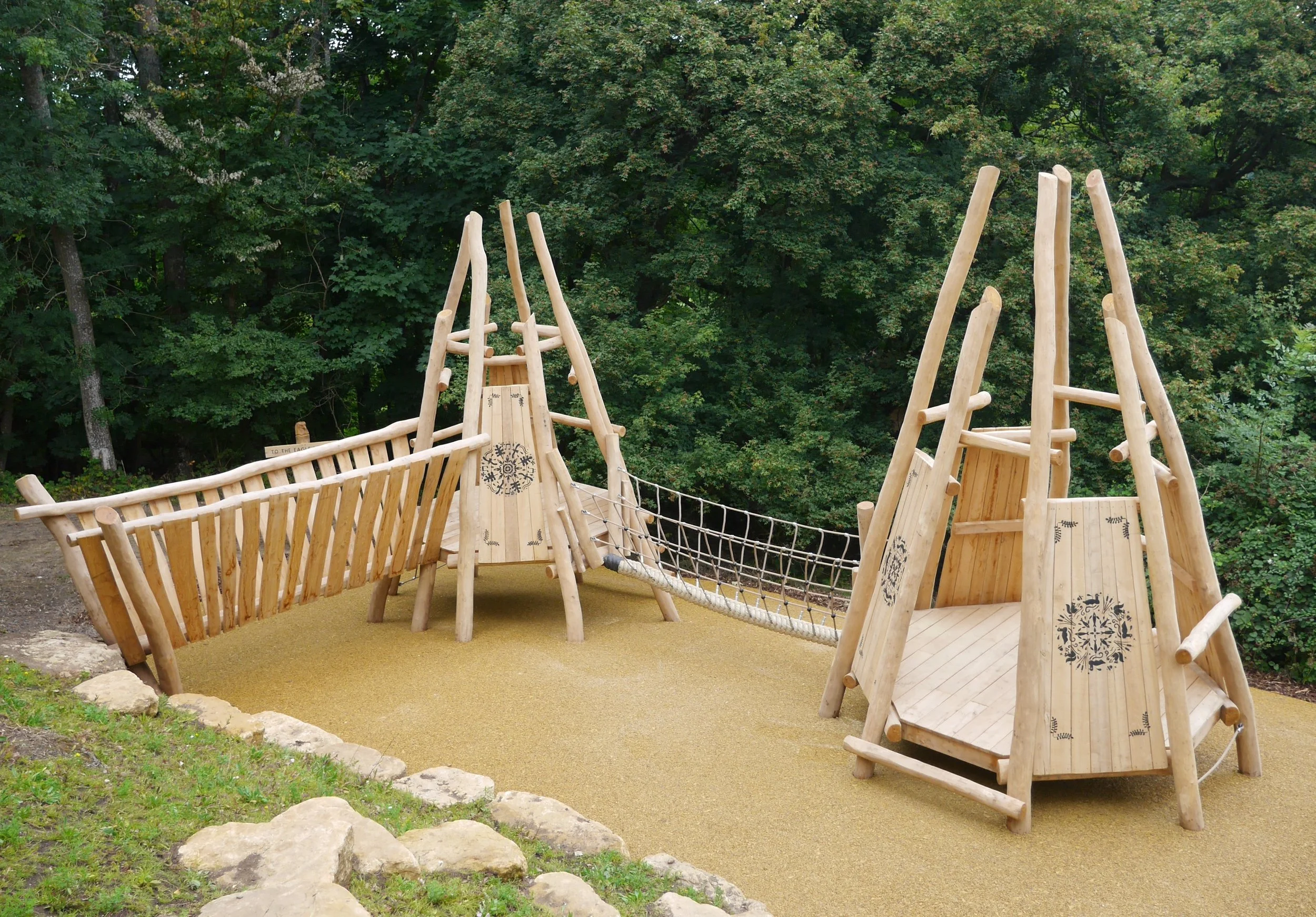Wooden playground structure with bridges, slides, and climbing areas, set on a sandy surface, surrounded by greenery and trees.