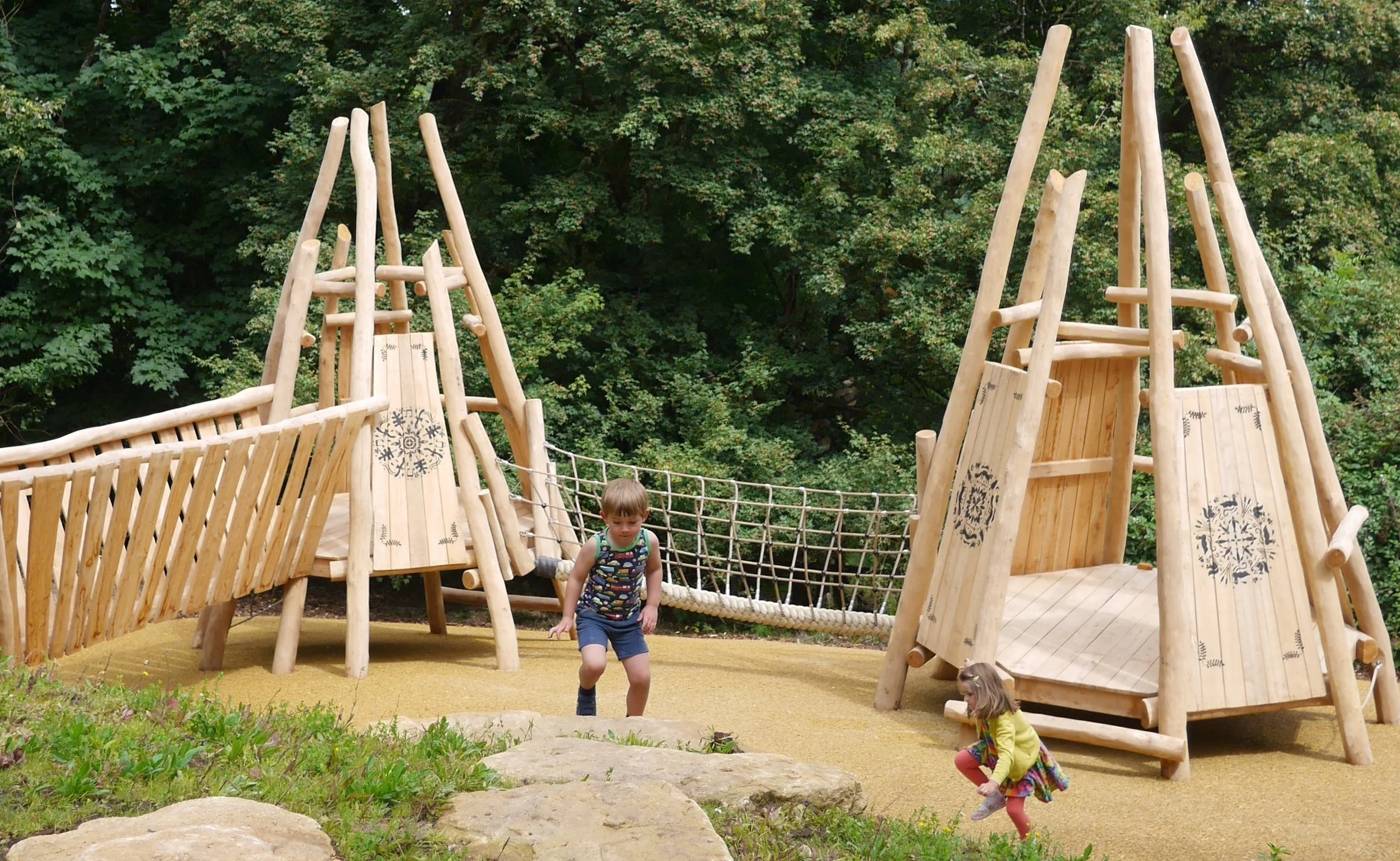 Two children playing on a wooden playground structure with a net bridge, jungle gym elements, and a slide, surrounded by greenery and trees.