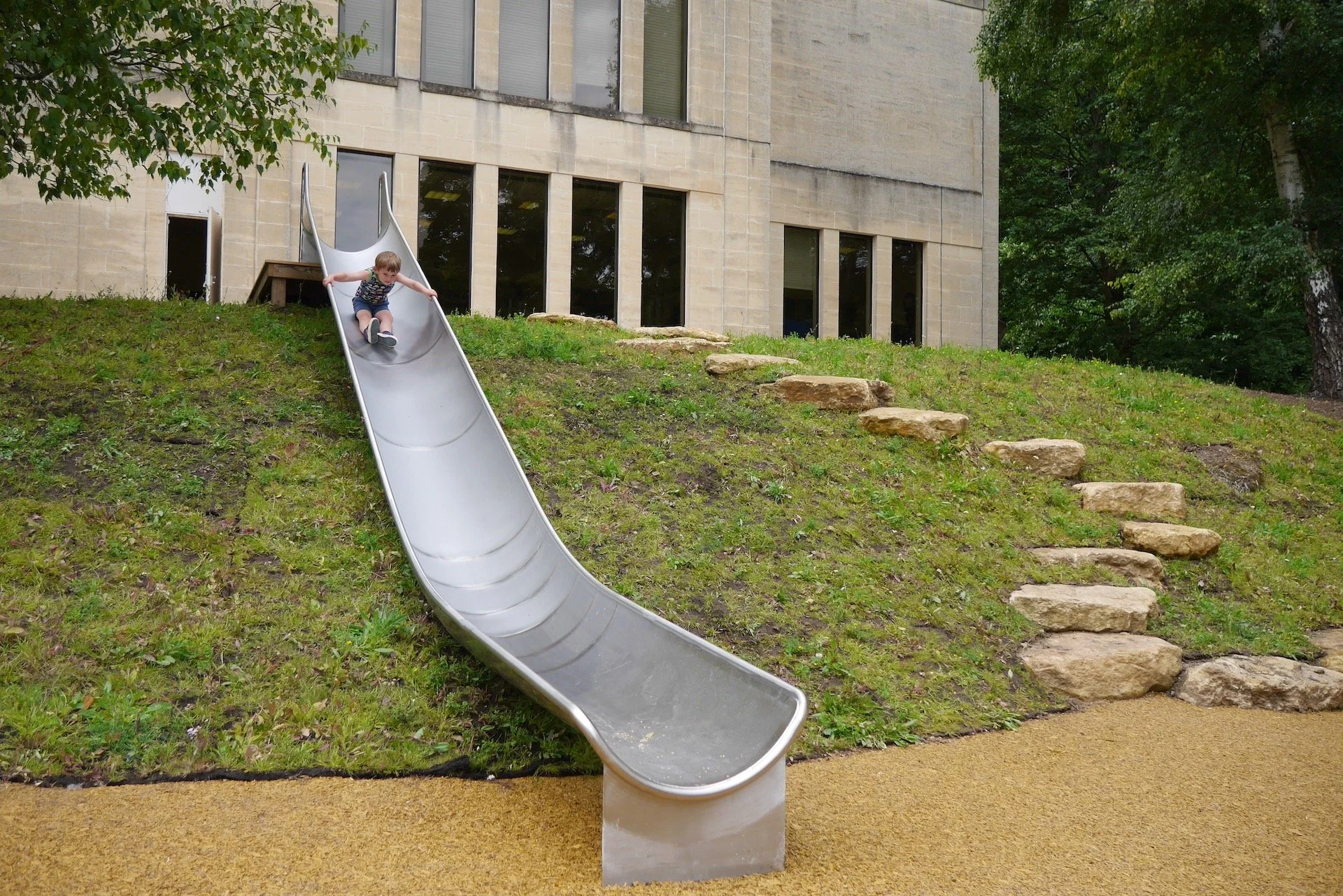 A child sliding down a metal slide on a grassy hill with stone steps on the side, next to a building with large windows and surrounded by trees.