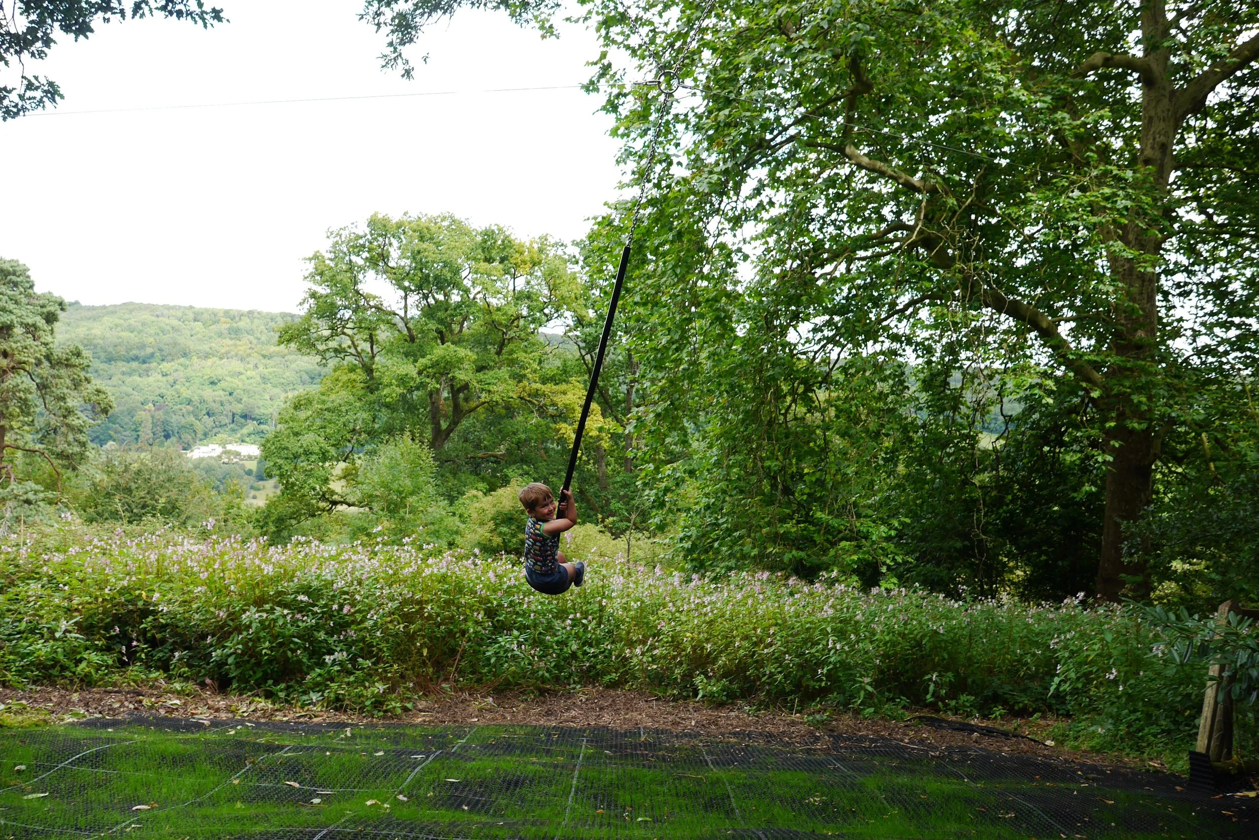 A young boy swinging on a tire swing outdoors in a lush, green park with trees and hills in the background.