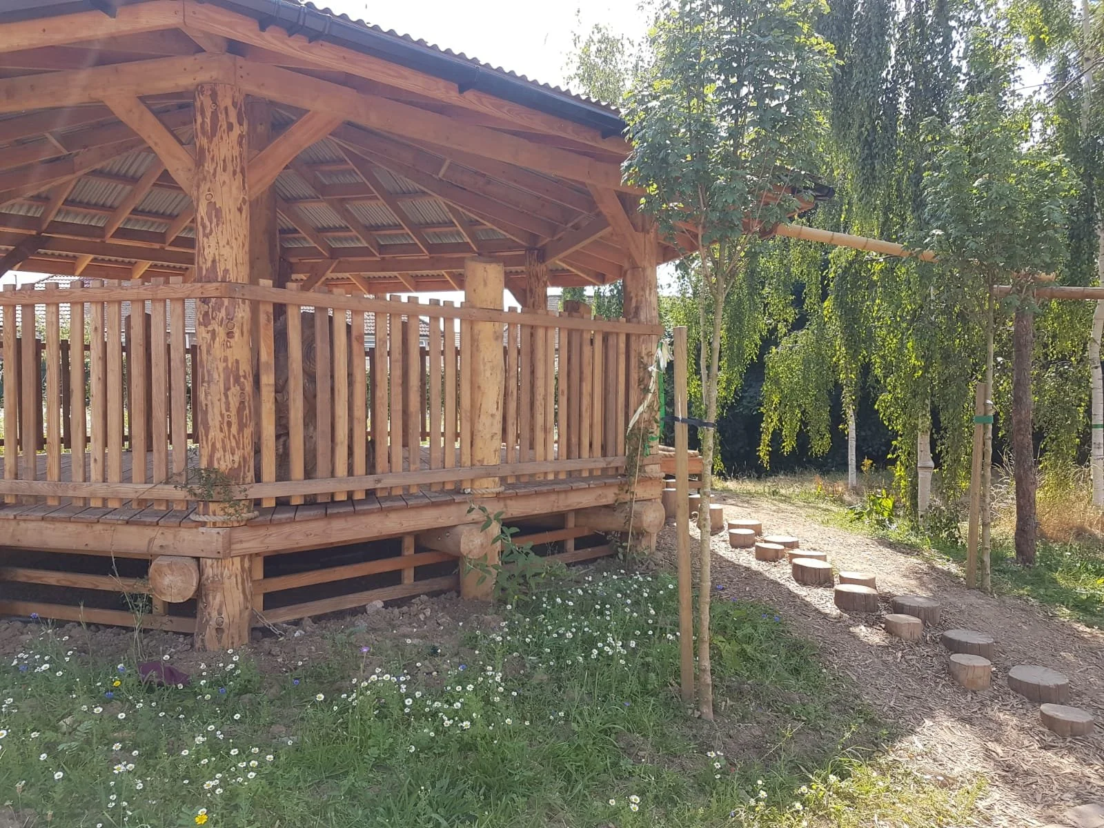 Wooden outdoor pavilion with a raised deck, surrounded by trees and flowers, with a stepping stone path leading to it.