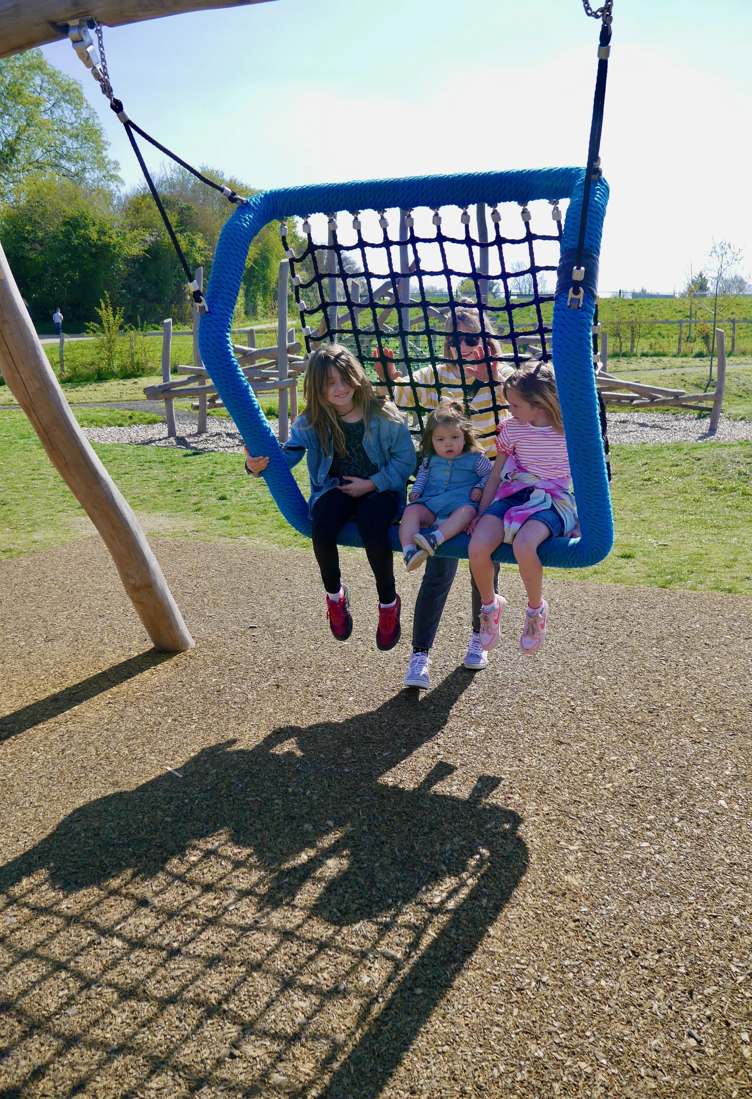 Four children sitting on a blue swing at a playground on a sunny day with trees and grass in the background.