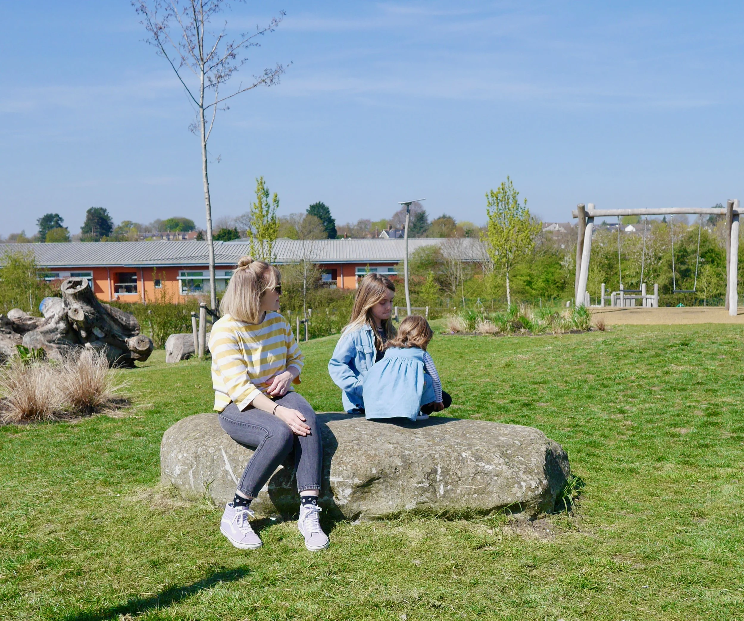 Three girls sitting on a large rock in a park on a sunny day, with trees, grass, a playground swing, and residential buildings in the background.