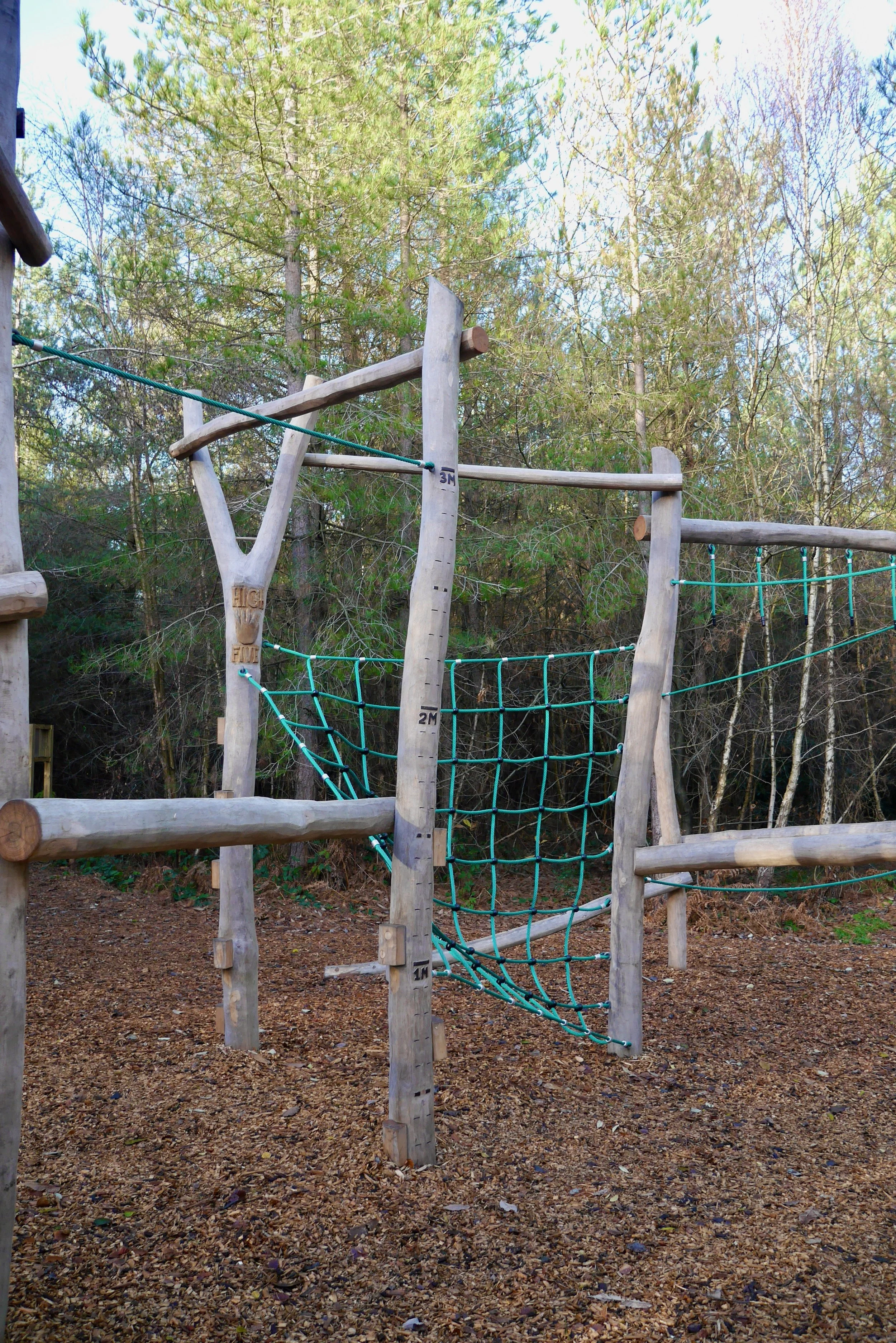 Wooden play structure with ropes and climbing features in a wooded area.