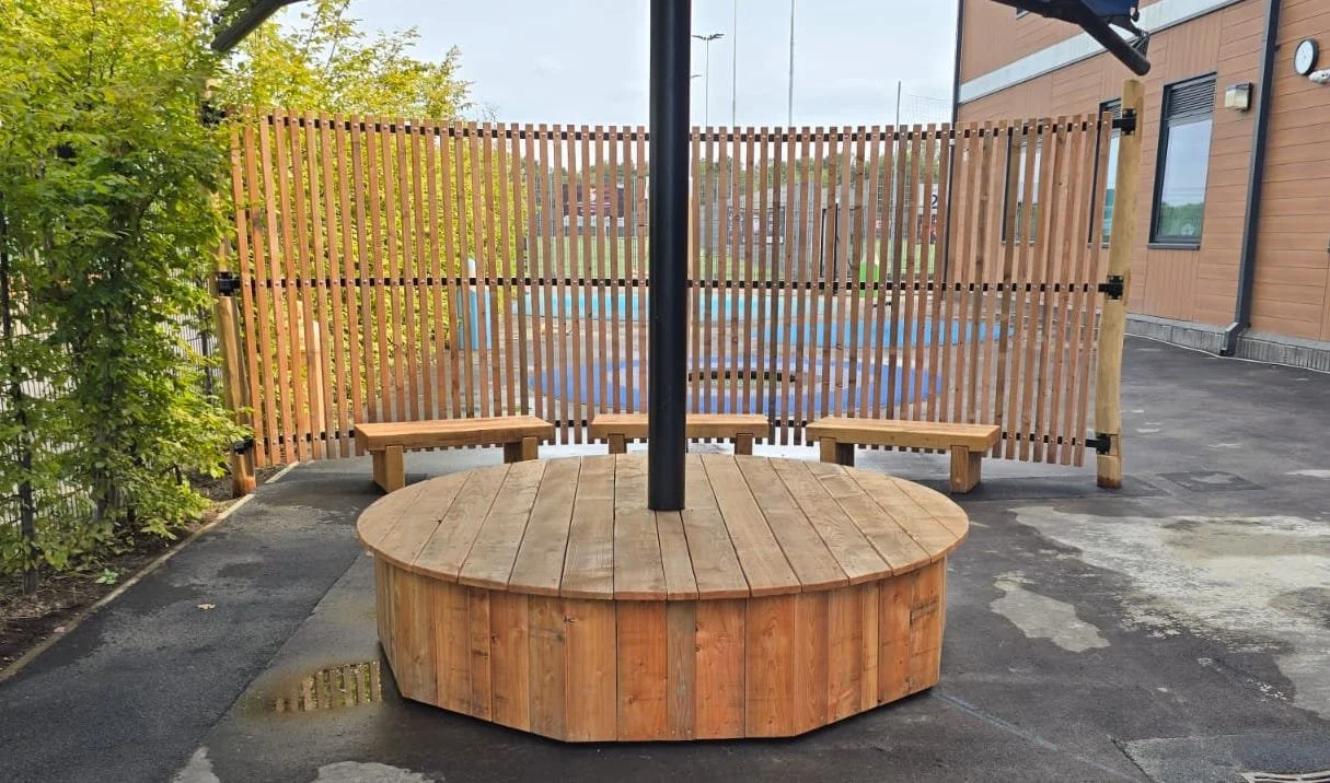 A wooden circular table with a black umbrella pole in the center, surrounded by three small wooden benches, located in an outdoor area with pavement, a wooden fence, and nearby building.