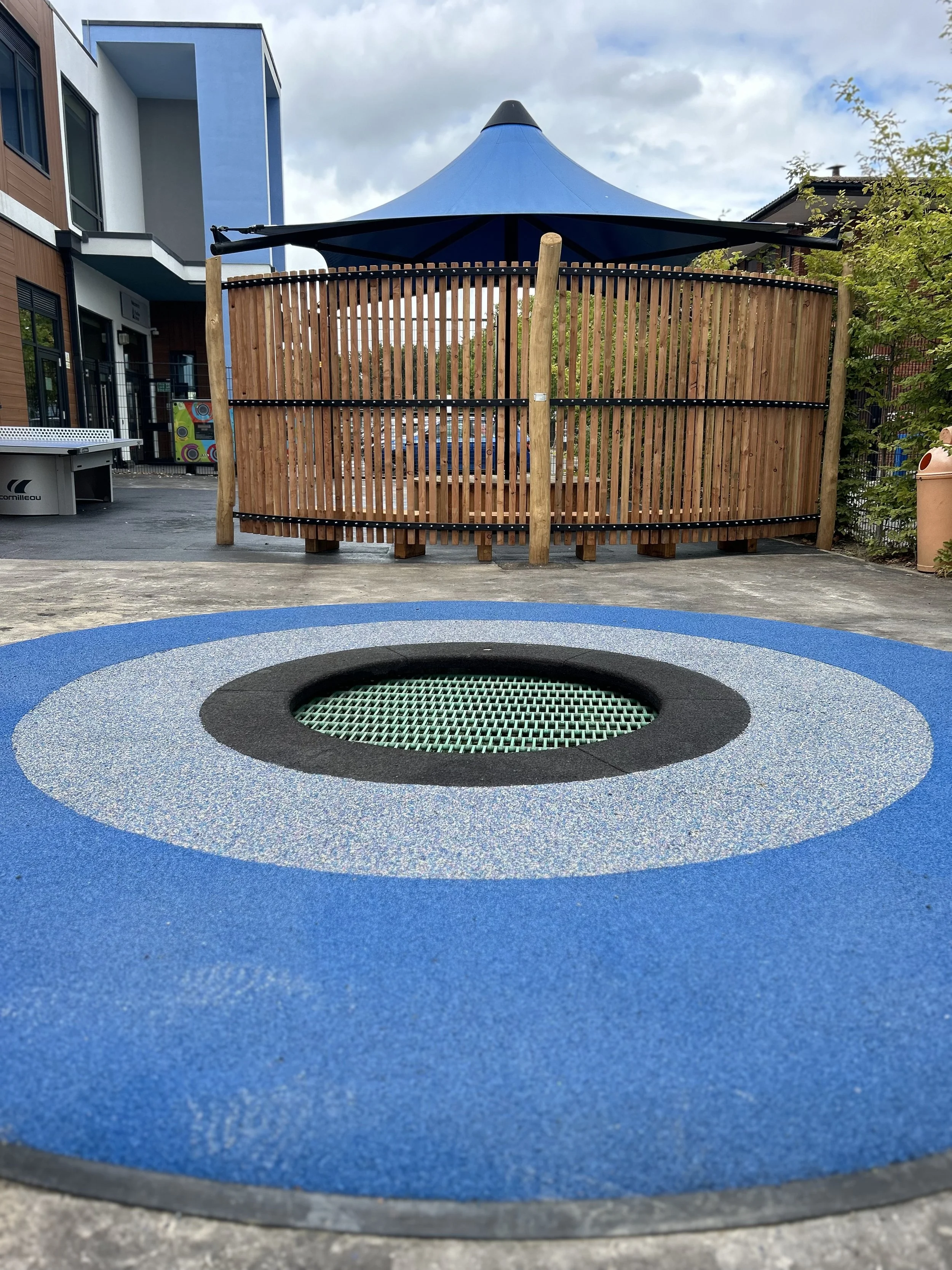 Blue trampoline with a circular black and green net in the center in a playground, with a wooden fence, shade structure, and nearby building in the background.