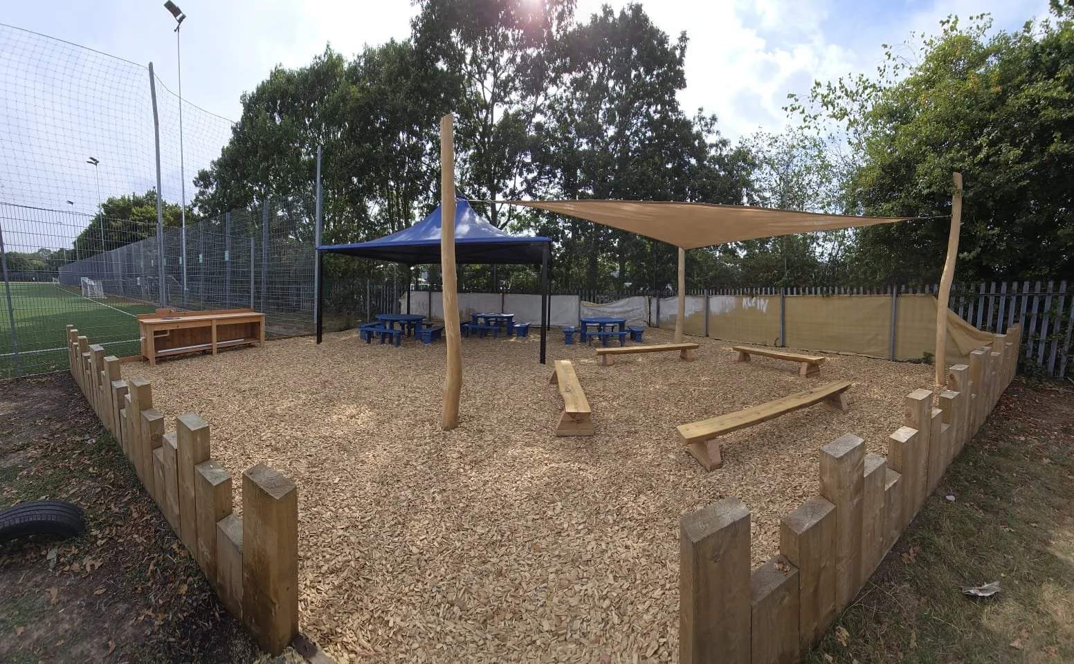 An outdoor children's playground area with wood chip ground cover, surrounded by a wooden fence. There are benches and a shade sail, with a blue canopy providing additional shade. Adjacent to a sports field with a tall fence, visible sports goal, and