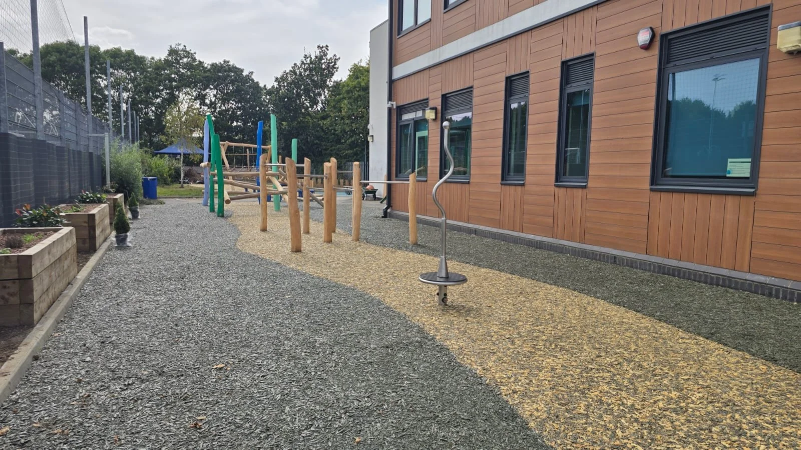 A playground area adjacent to a building with wooden siding, featuring a curved metal spinning toy, wooden climbing structures, and planter boxes along the left side, with a gravel and yellow-yellow textured path.