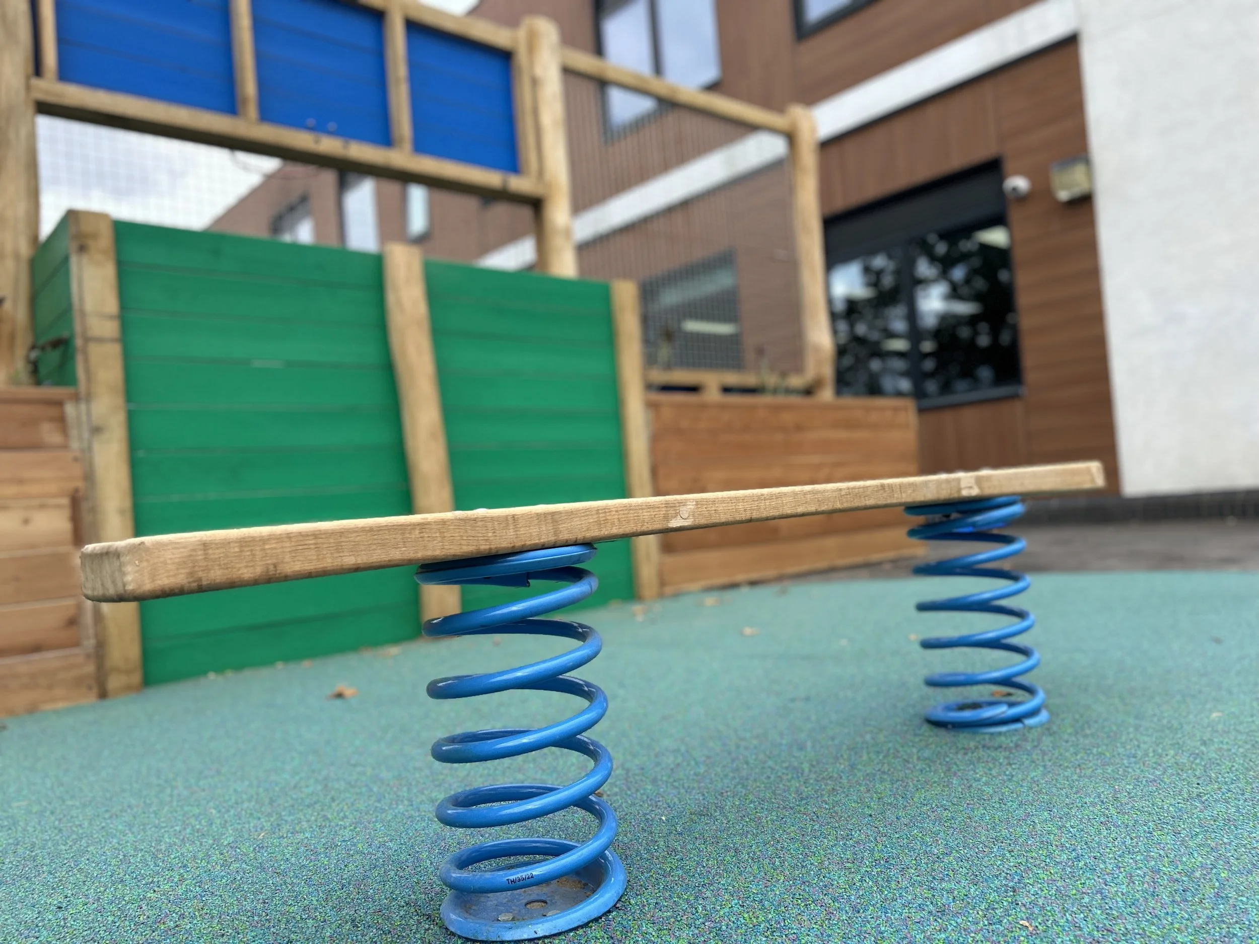 Close-up of a small wooden spring rocker on a colorful rubber playground surface, with a wooden play structure and a building with large windows in the background.