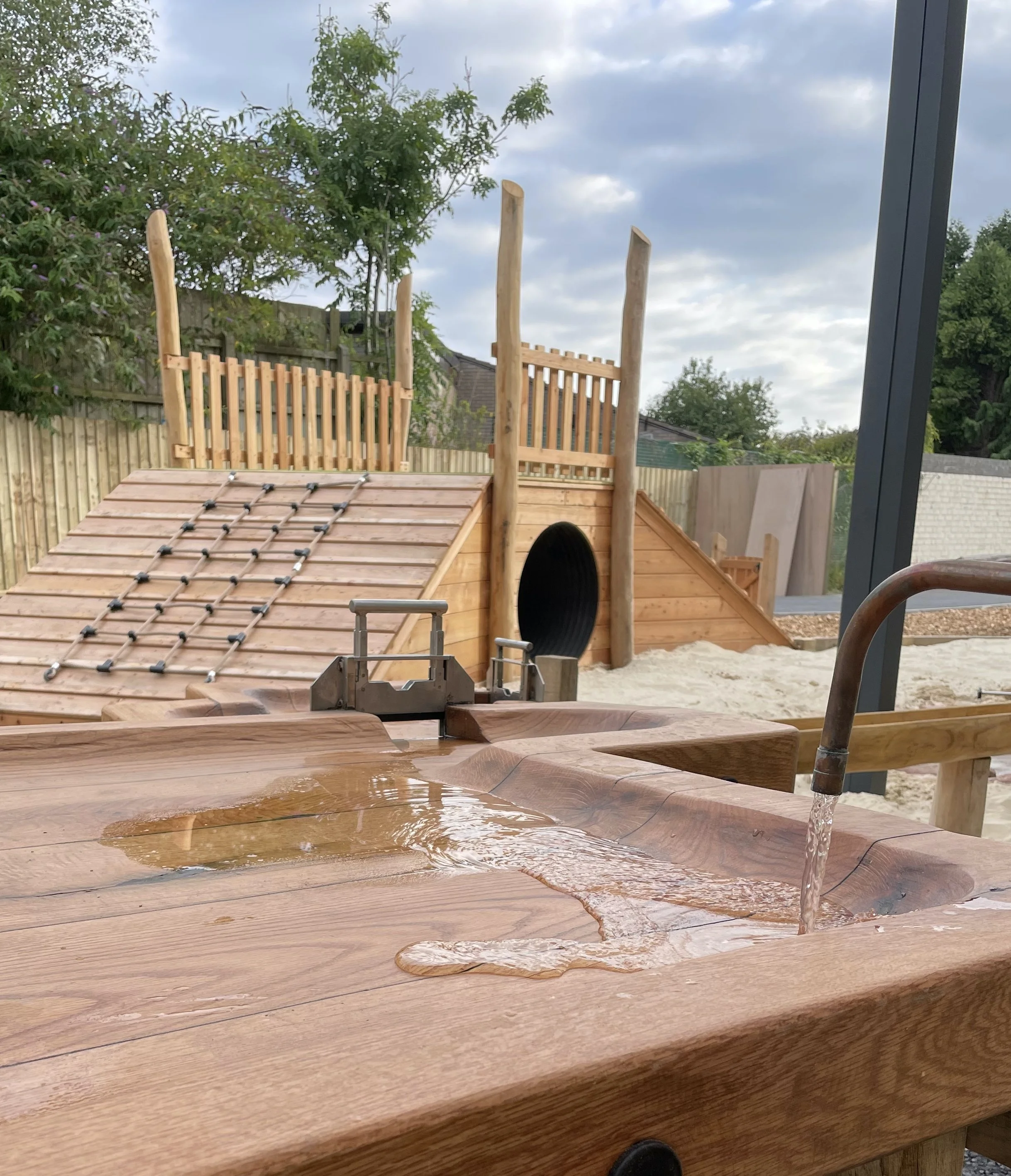 A wooden outdoor play area with a sandbox, a climbing structure with a slide, and a water feature with flowing water, set against a background of trees and a cloudy sky.