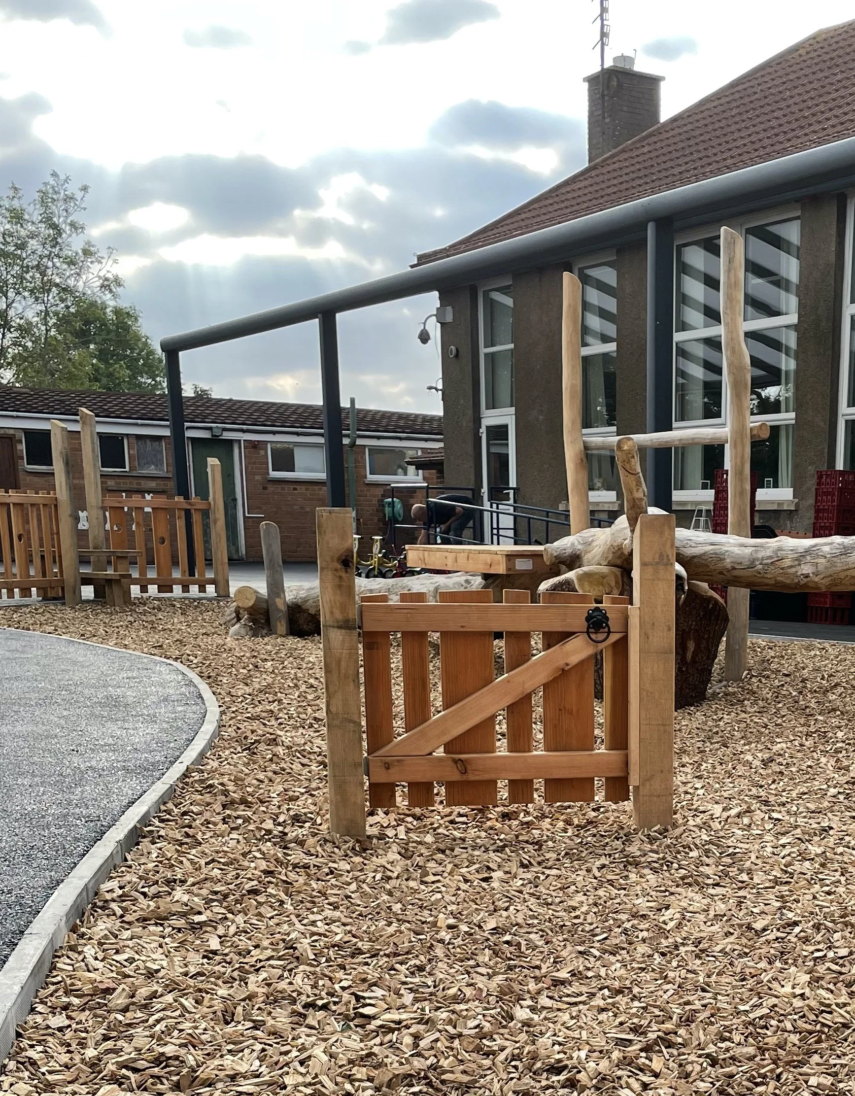 A playground area with wood chip ground cover and a small wooden gate in the foreground. There are wooden play structures and a ramp with black handrails near a building with large windows and a chimney. The sky is partly cloudy with sunlight peeking