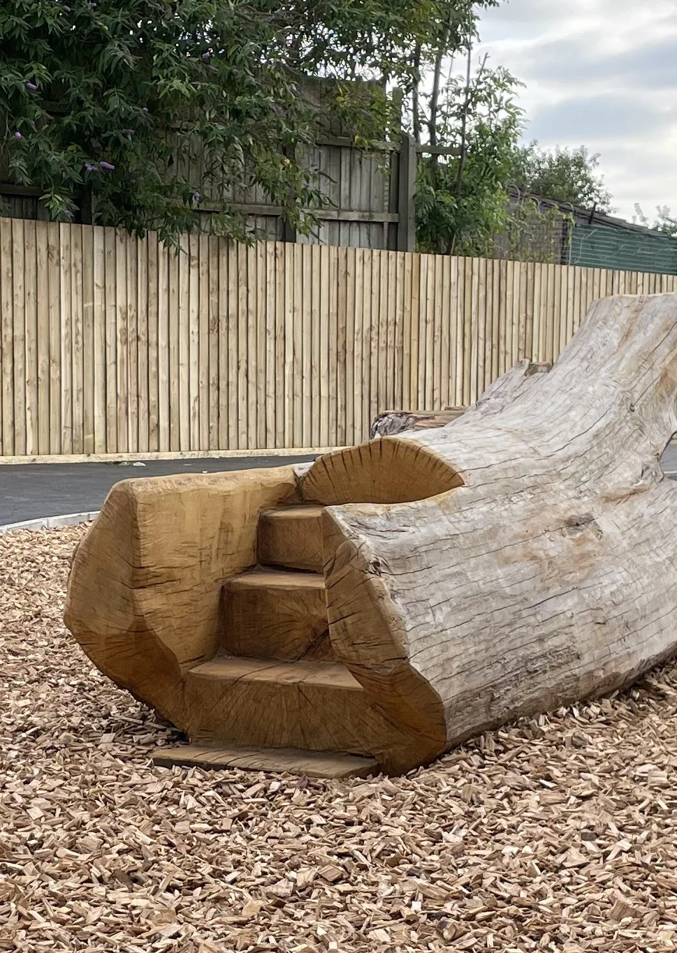 A large wooden sculpture resembling a seated animal, possibly a bear, with carved stairs in the middle, placed on a surface covered with wood chips in an outdoor setting.