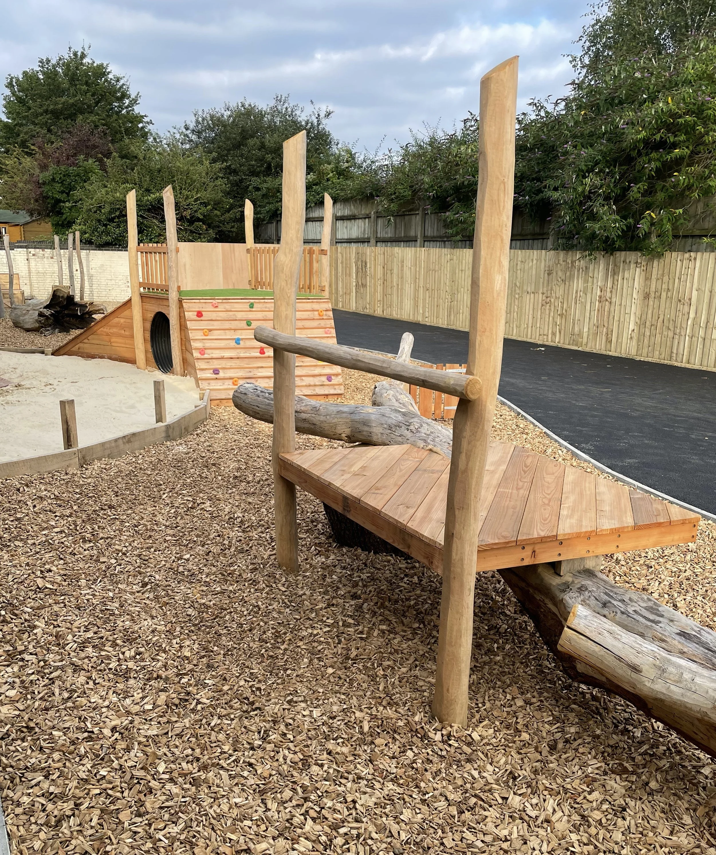 Playground with a wooden platform, logs, climbing wall, and sandy area, surrounded by a wooden fence and trees.