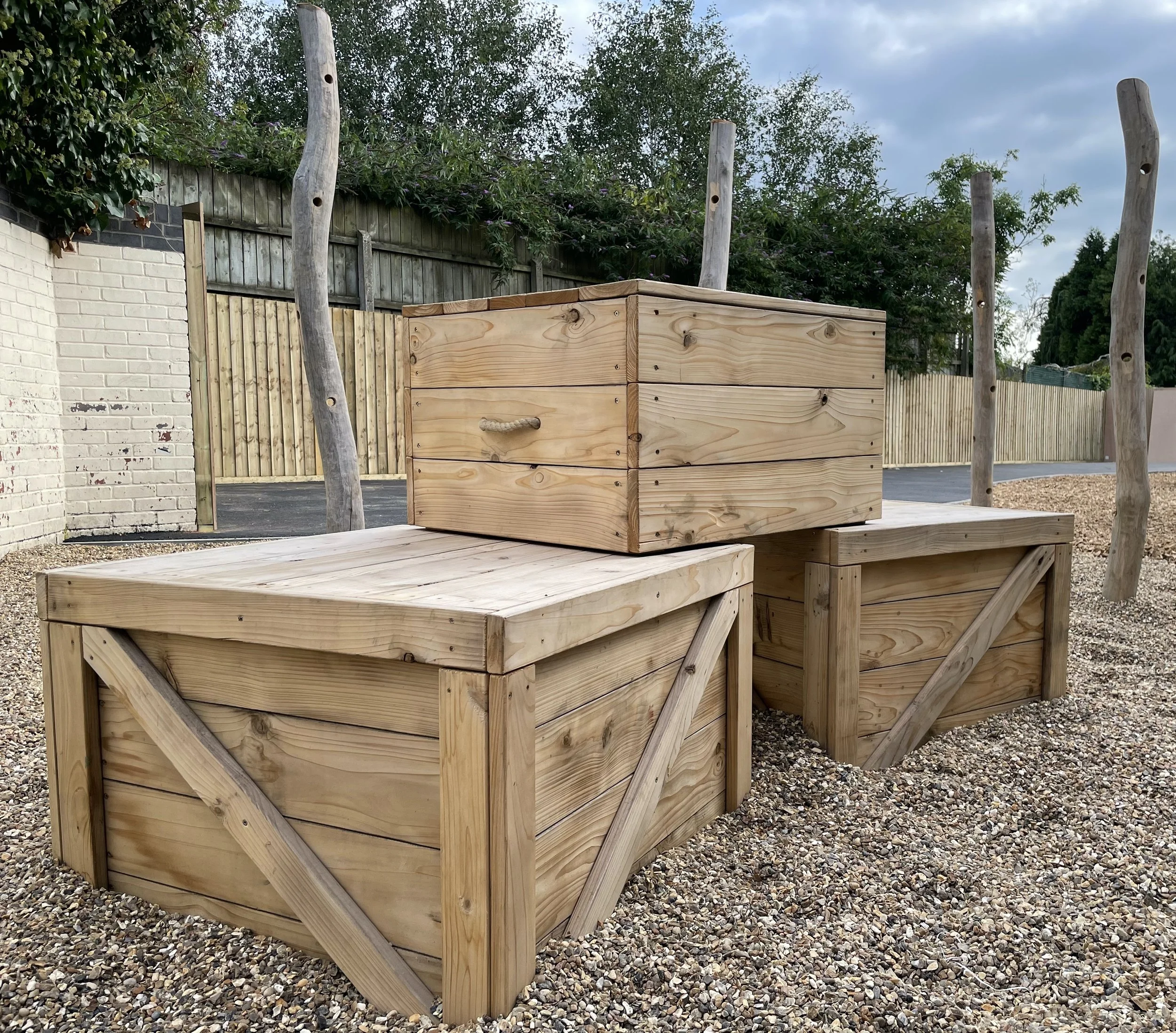 Three wooden storage boxes in an outdoor area with gravel ground and wooden posts, nearby brick wall, and trees in the background.