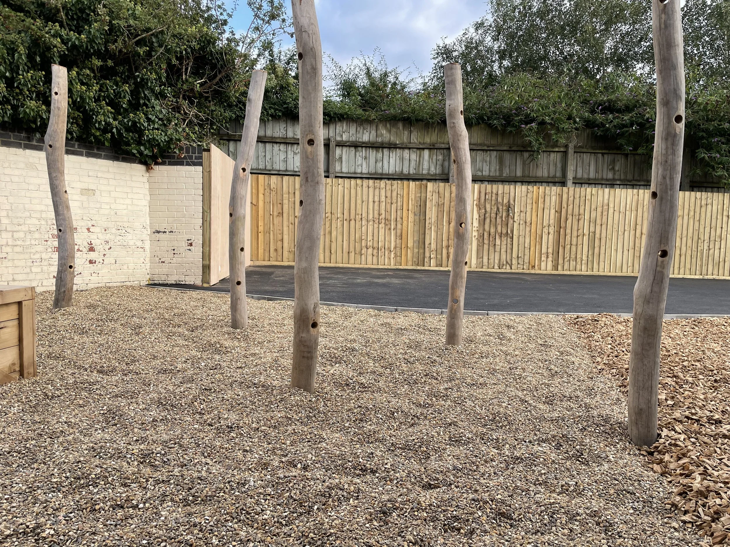 Outdoor playground area with five tall wooden poles with holes, gravel ground, a paved section in the background, and wooden fencing along the perimeter.