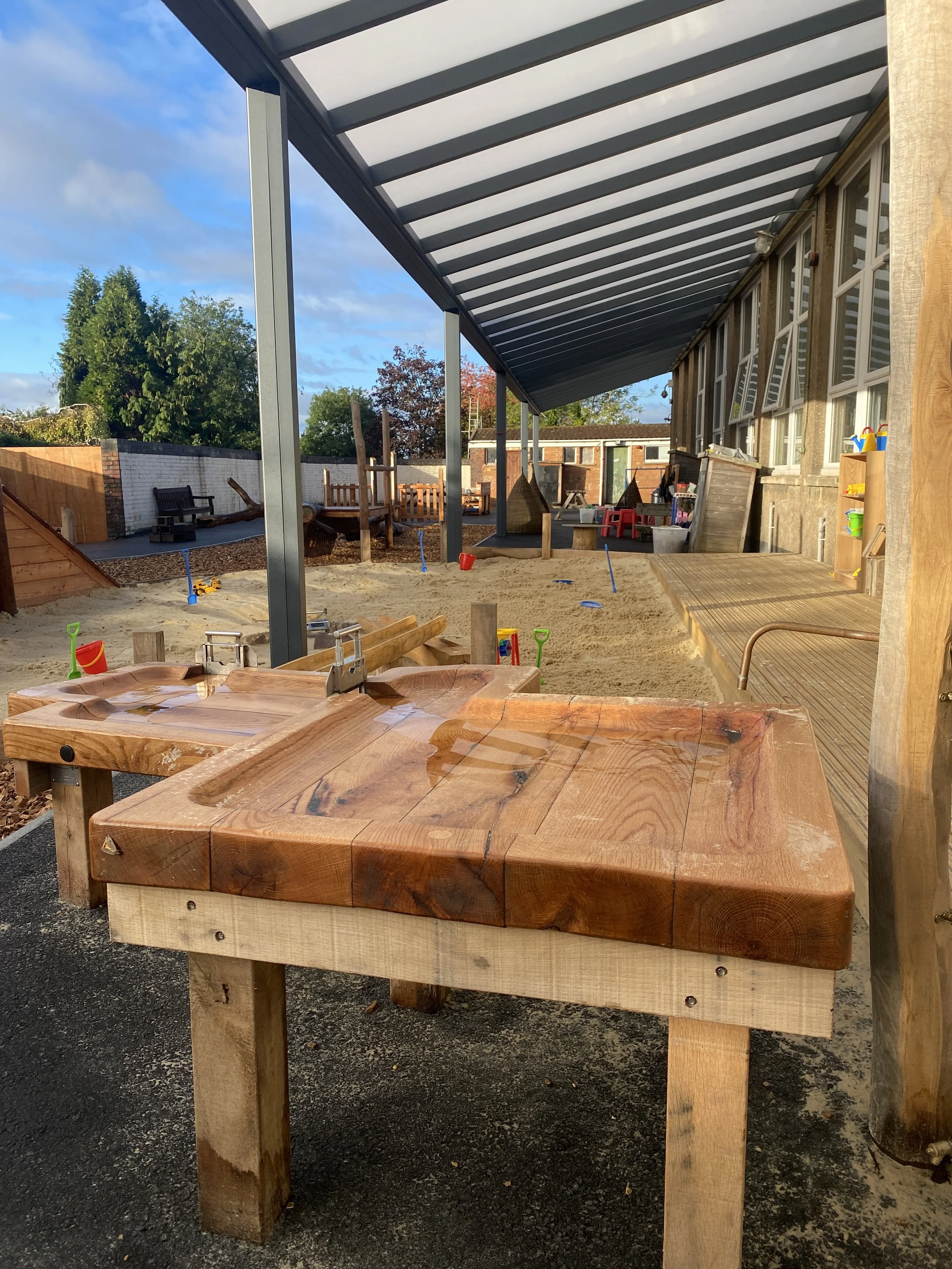 Children's outdoor sandbox with wooden water table and sand toys, covered patio with windows, and playground equipment in the background.