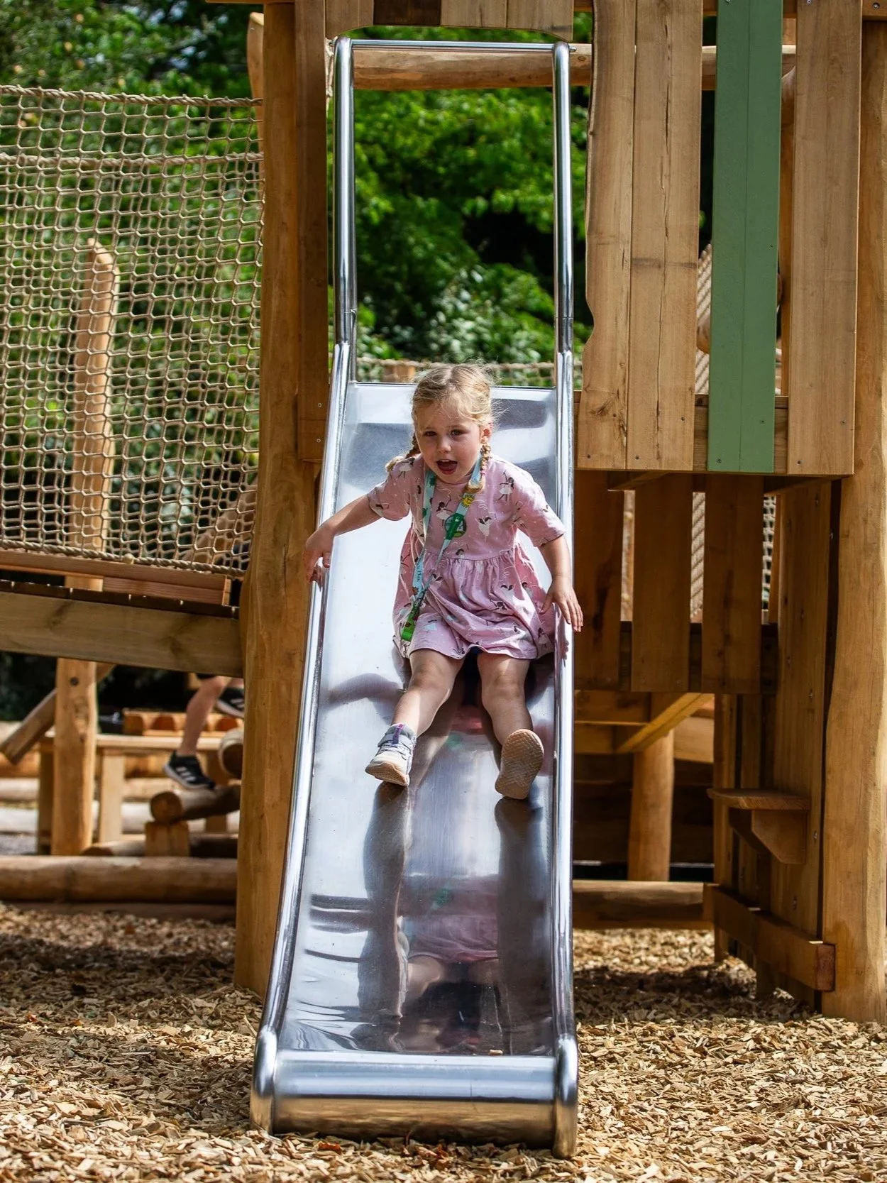 A young girl in a pink dress sliding down a metal slide at a wooden playground surrounded by wood chips and green trees.