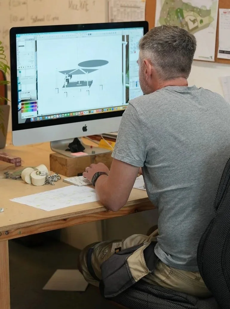 A man working at a desk with a large iMac computer, designing a 3D structure. The desk has papers, tape, and keychains. Behind him, a corkboard displays maps, plans, and notes on a wall.