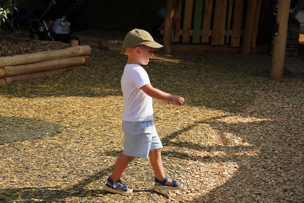 Young boy walking outdoors on a gravel surface, wearing a tan hat, white t-shirt, light-colored shorts, and sandals, with trees and a wooden structure in the background.