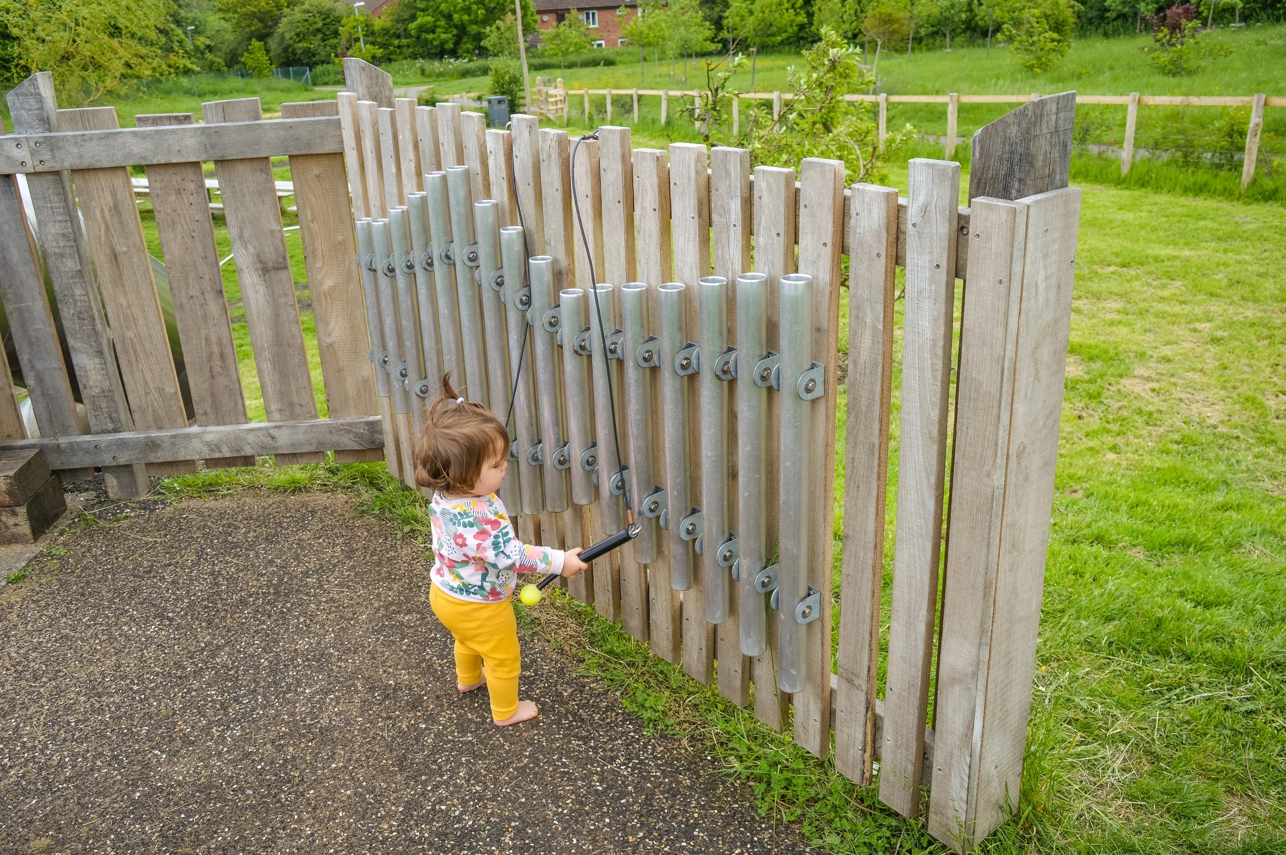 A young girl playing a xylophone with large metallic tubes outdoors, standing on a textured ground near a wooden fence with a grassy field and trees in the background.