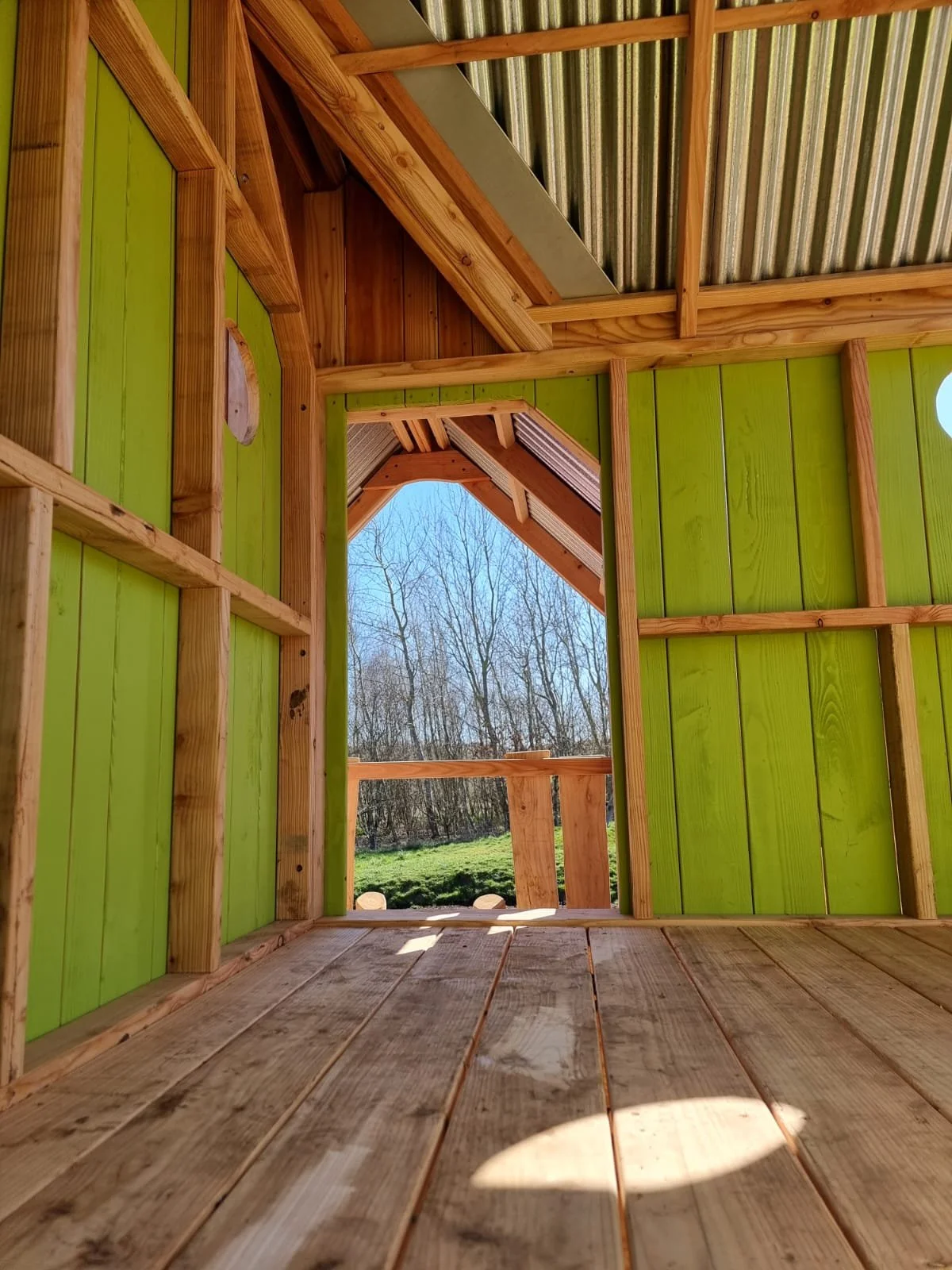 Interior of a wooden structure under construction with green painted walls, exposed framing, and a view of trees and grass outside through an opening.