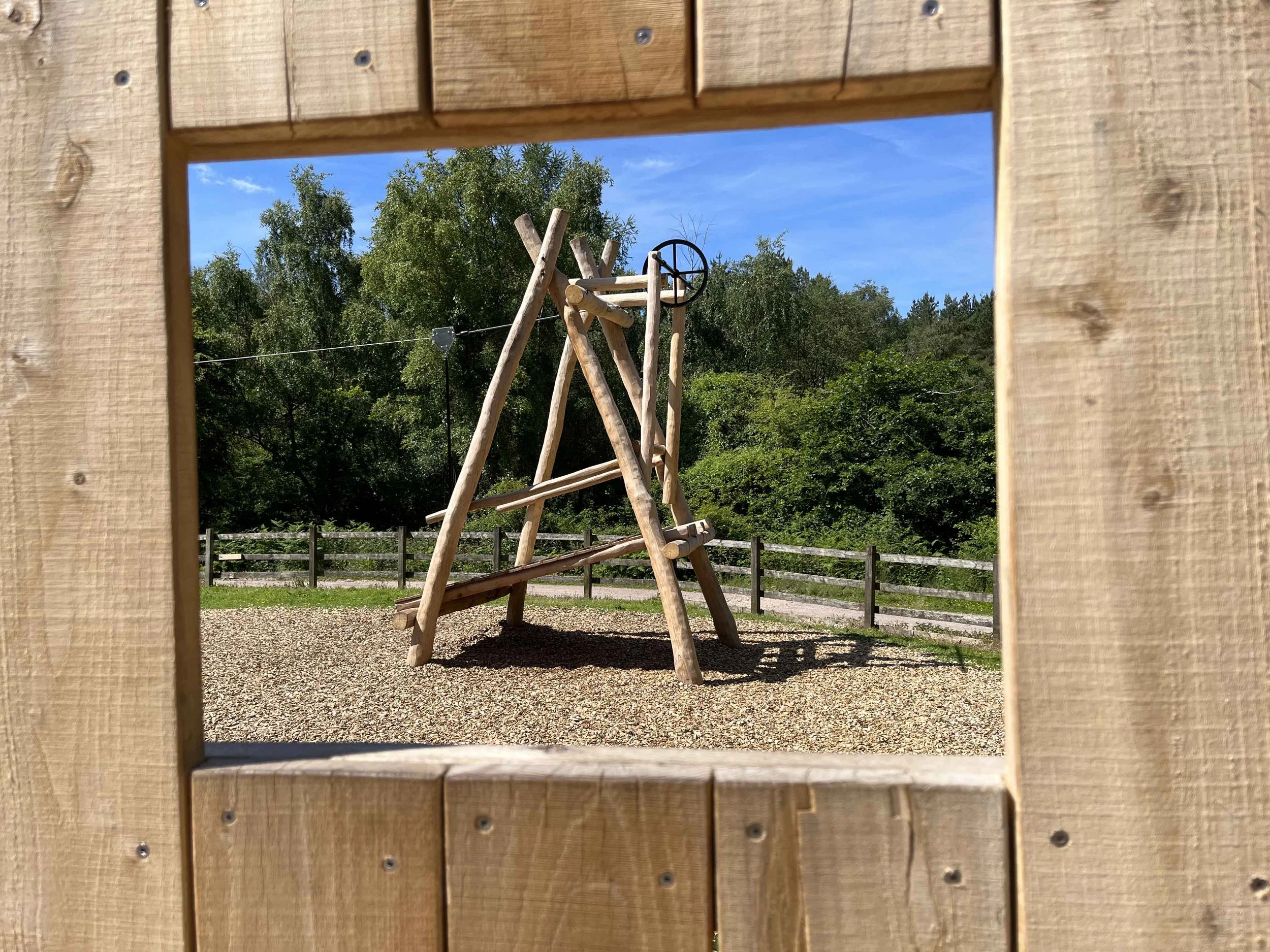 View of a wooden playground structure through a square cutout, with trees and a blue sky in the background.