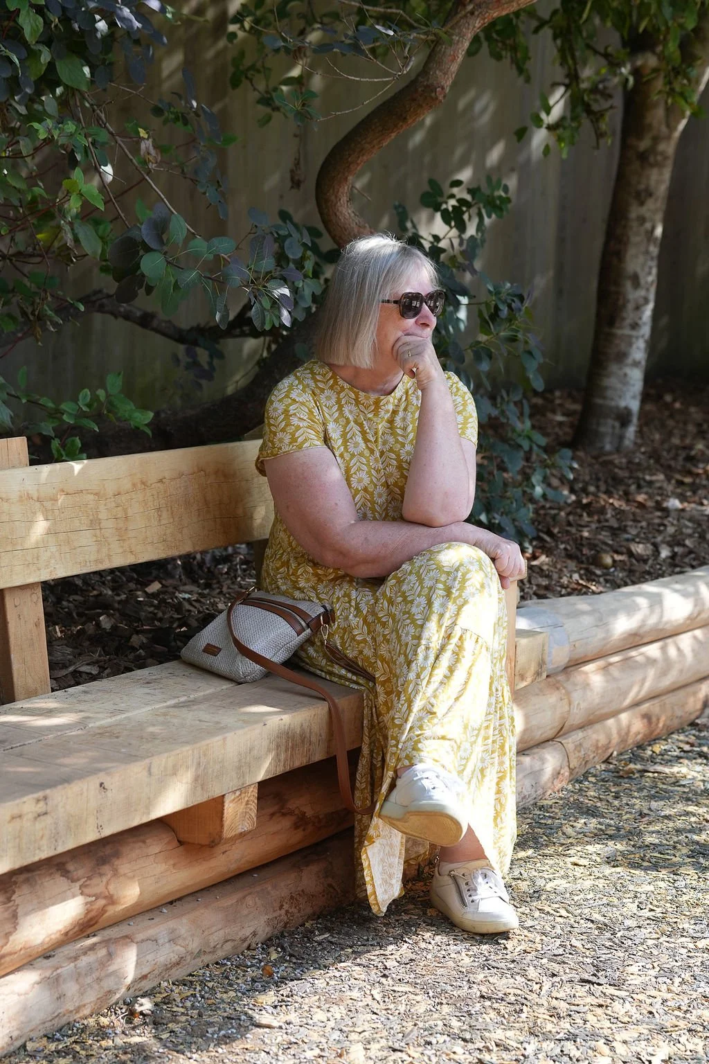 An older woman with gray hair, wearing sunglasses and a yellow floral dress, is sitting on a wooden park bench, resting her chin on her hand, appearing thoughtful.