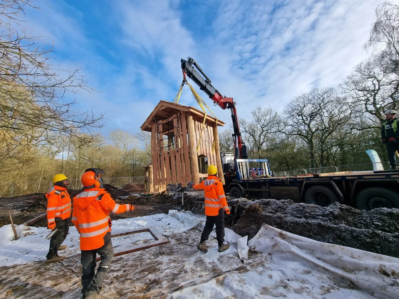 Construction workers in orange safety vests and yellow helmets operate a crane lifting a small wooden house structure at a construction site surrounded by trees.