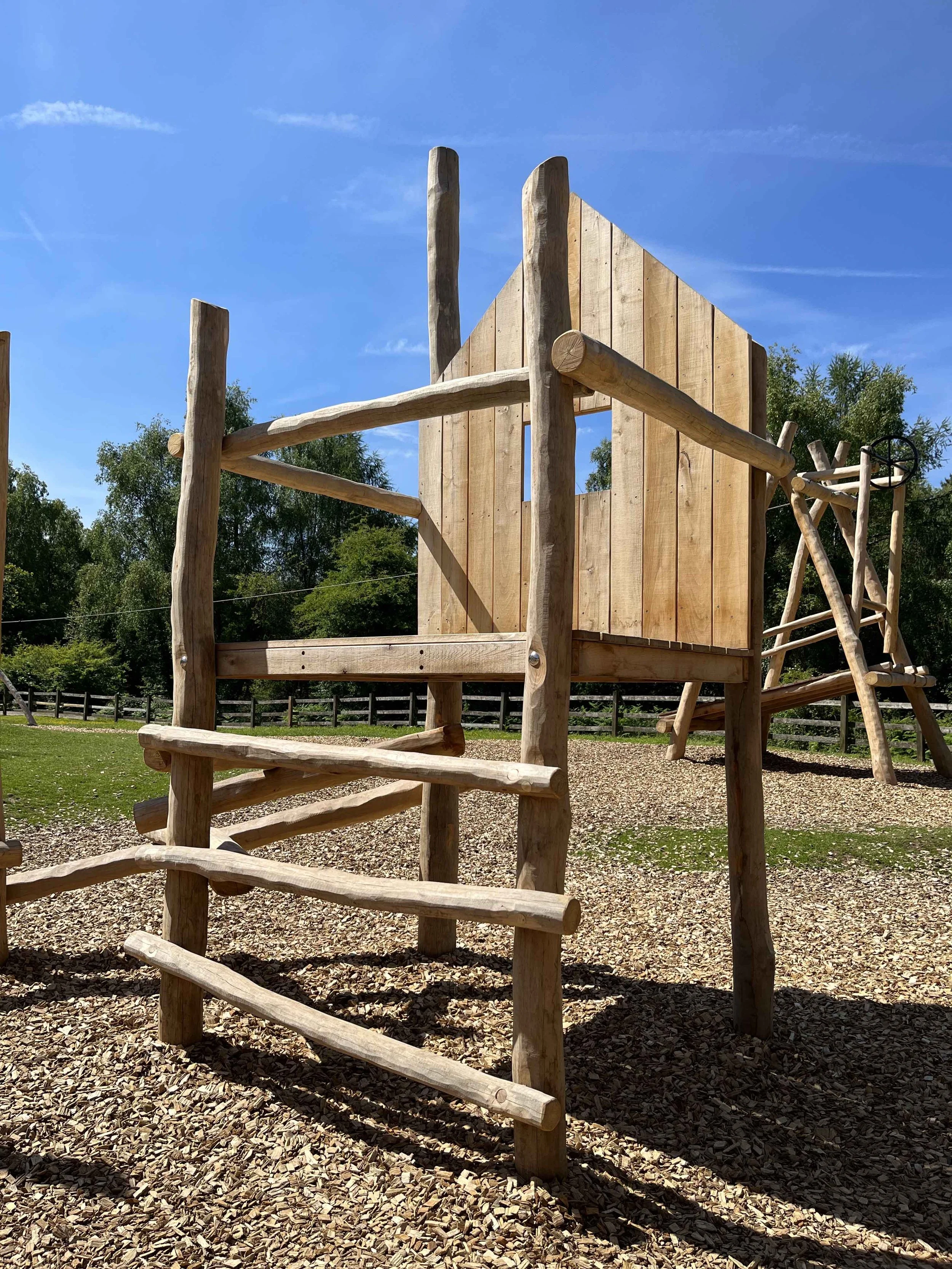 Wooden playground structure with climbing ladder and slide, set in a park under a blue sky with some clouds.